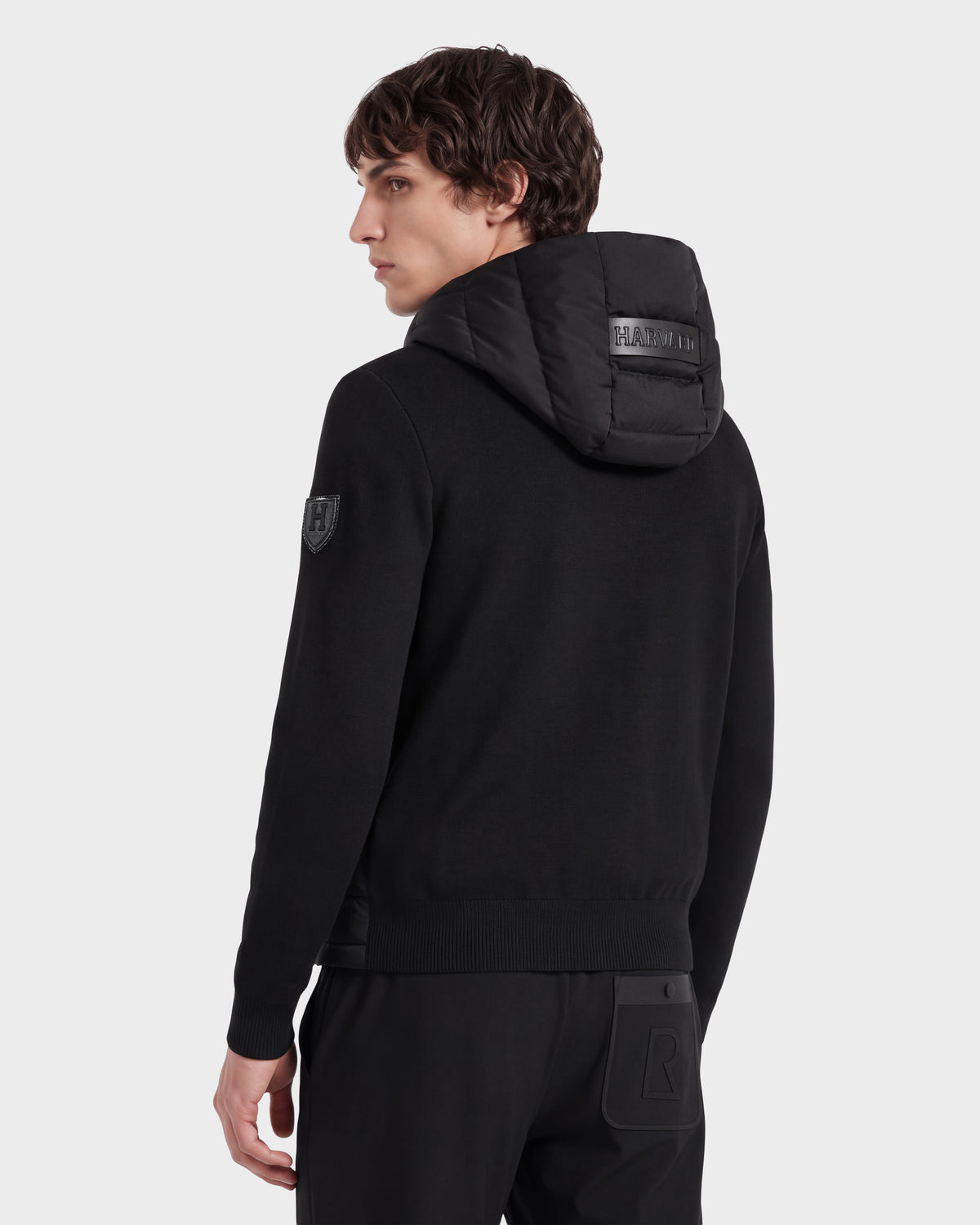 A person with short brown hair is seen from behind against a plain background, wearing the Rudsak RIVER HV - H125503 Black hooded sweatshirt featuring a patch on the sleeve and raised lettering on the hood, giving the outfit an exclusive, limited edition appeal.