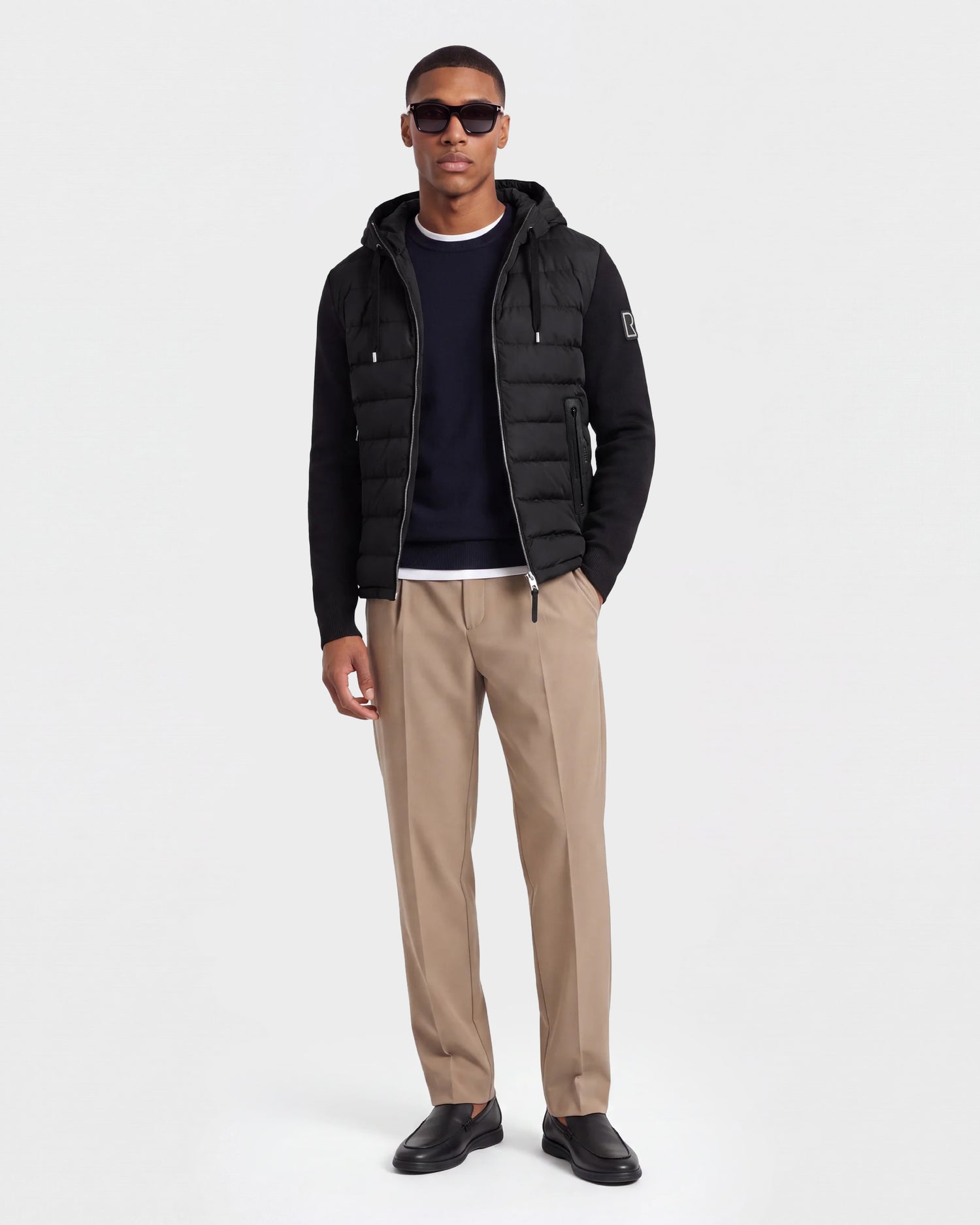 A man wearing sunglasses and the RIVER - 6124584 Black hooded down jacket by Rudsak, along with a black sweater featuring a white collar, beige trousers, and black shoes, stands against a plain white background.