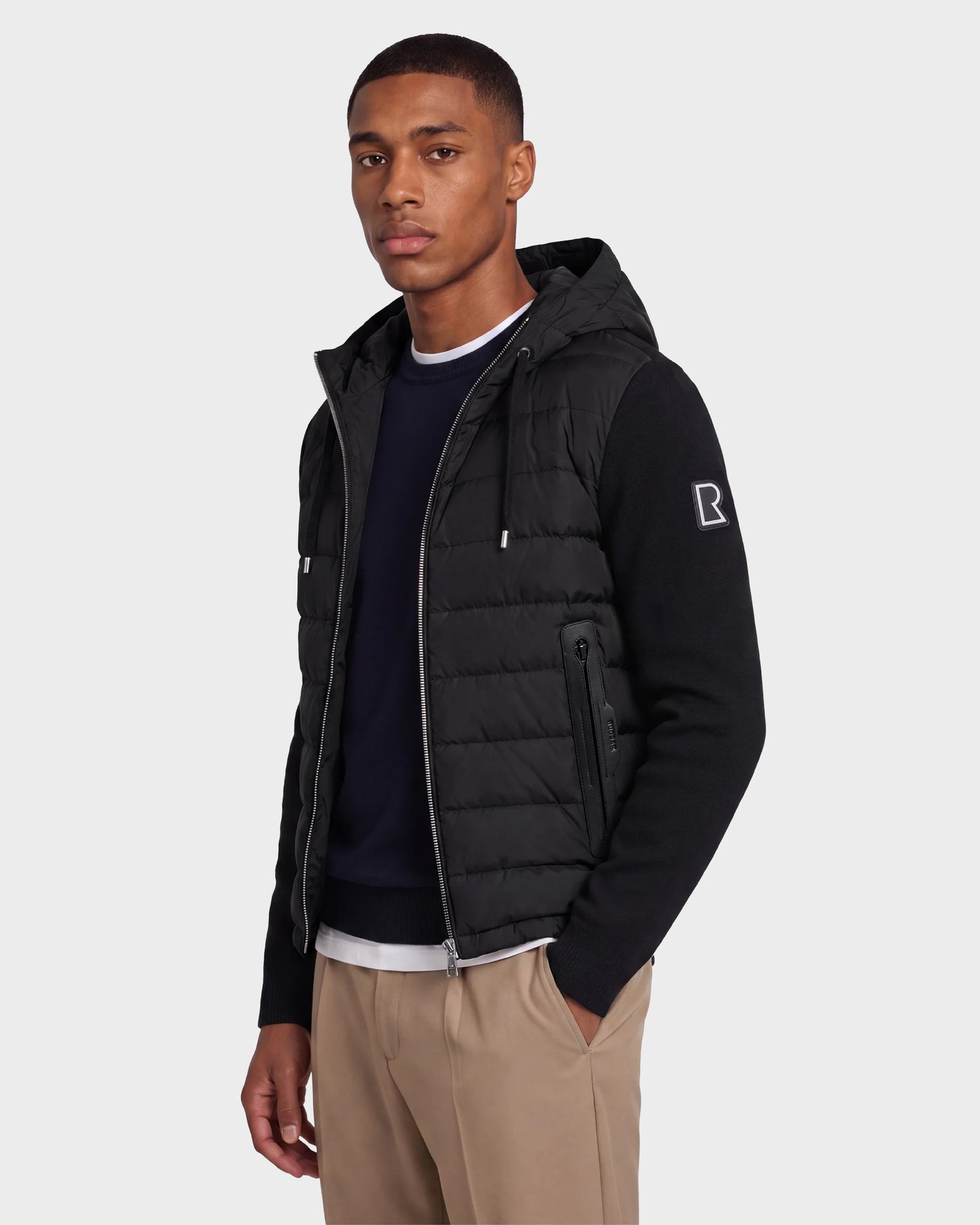 A man wearing the Rudsak RIVER - 6124584 black hooded down jacket with a B patch on the sleeve, a navy sweater, and tan pants stands against a light gray background.