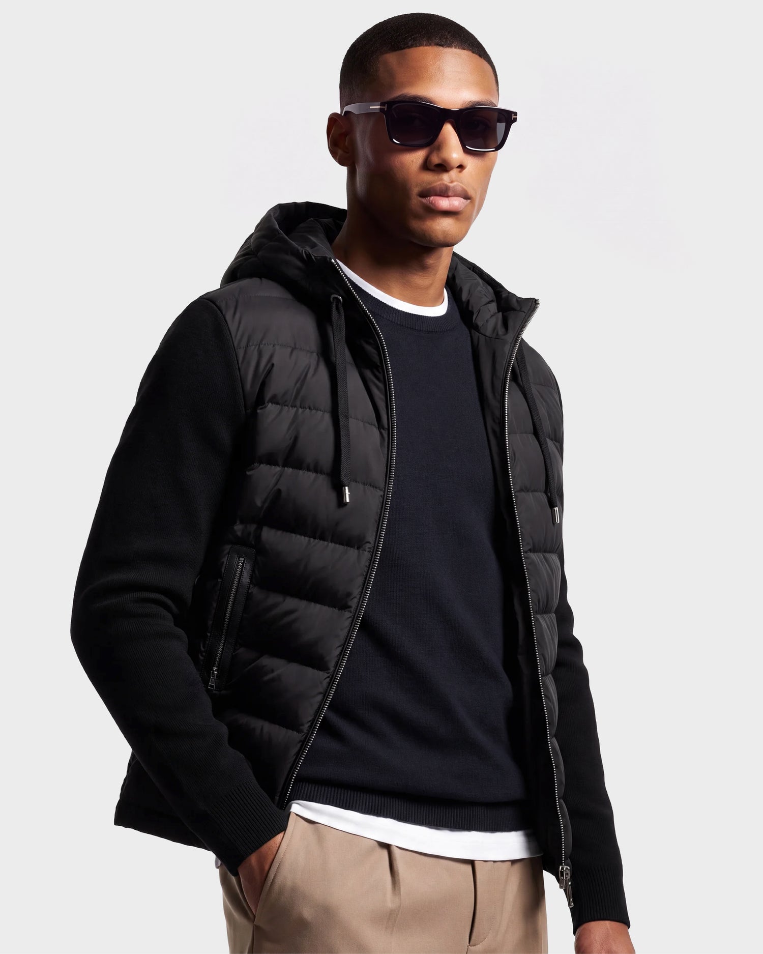 A man stands against a plain backdrop with one hand in his pocket, wearing the Rudsak RIVER - 6124584 Black hooded down jacket layered over a black sweater and white shirt, paired with beige pants and sunglasses.
