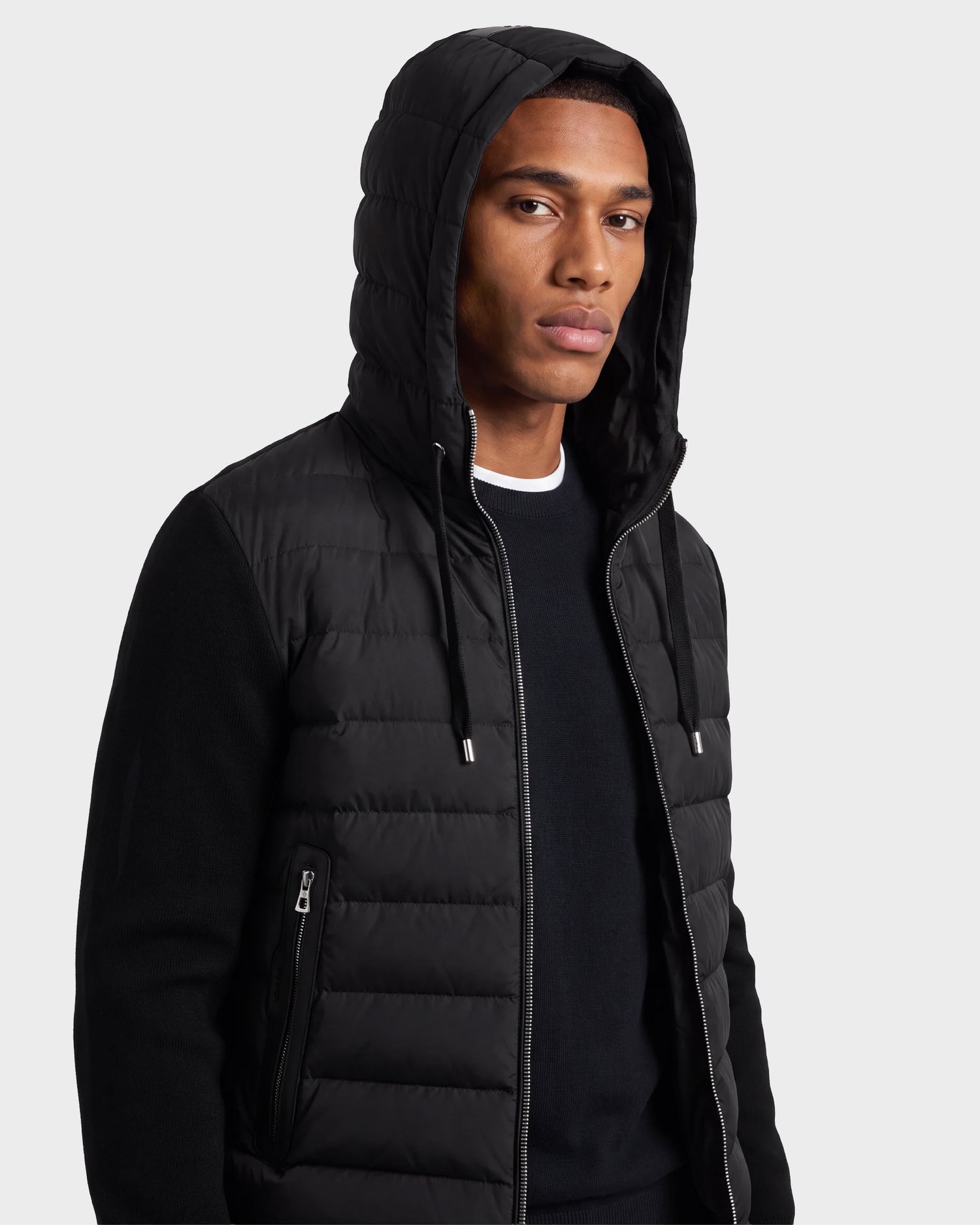 A man wearing the Rudsak RIVER - 6124584 Black quilted hooded down jacket over a black sweater, looking at the camera against a plain light gray background.
