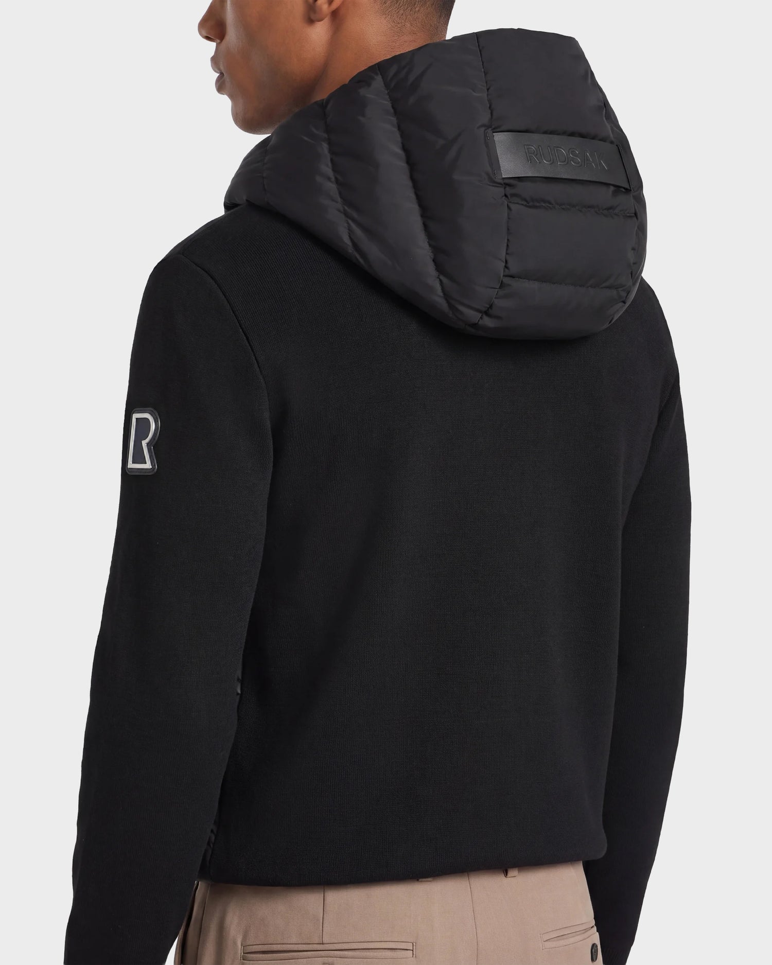 A man shown from the back wears the Rudsak RIVER - 6124584 Black hooded down jacket, featuring a padded hood with textured detailing and a sleeve patch. The word "BOSS" is embossed on the hood. He is also wearing beige pants.