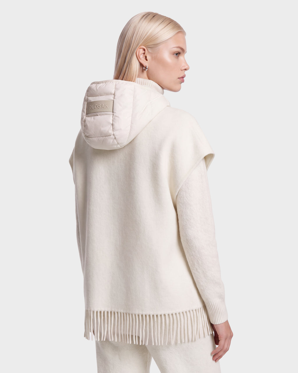 A woman with straight blonde hair wears the ROXANE - 8526677 Vanilla cream hooded poncho from Rudsak, featuring fringe detail at the hem and matching pants. A small label accents the hood as she faces away, gazing left against a light gray background.