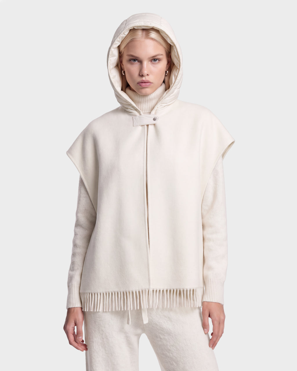 A woman stands facing forward against a plain light background, wearing the ROXANE - 8526677 Vanilla hooded cape with fringe at the hem by Rudsak, paired with a matching sweater and pants.