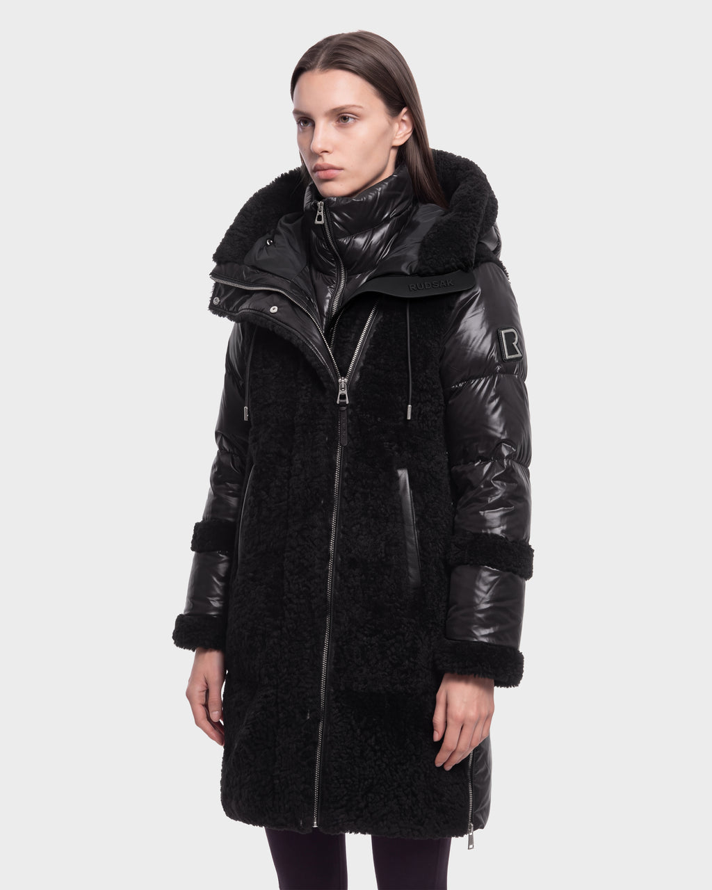 A woman stands facing forward with a neutral expression against a plain white background, wearing the Rudsak SHELLEY - 8126729 Black winter coat, which is long, black, shiny, and textured with a high collar and hood.