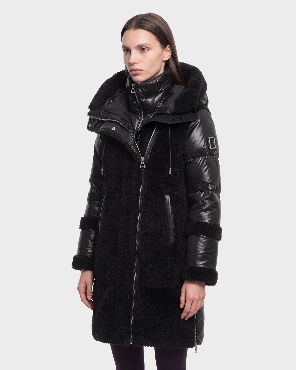 A woman stands facing forward with a neutral expression against a plain white background, wearing the Rudsak SHELLEY - 8126729 Black winter coat, which is long, black, shiny, and textured with a high collar and hood.