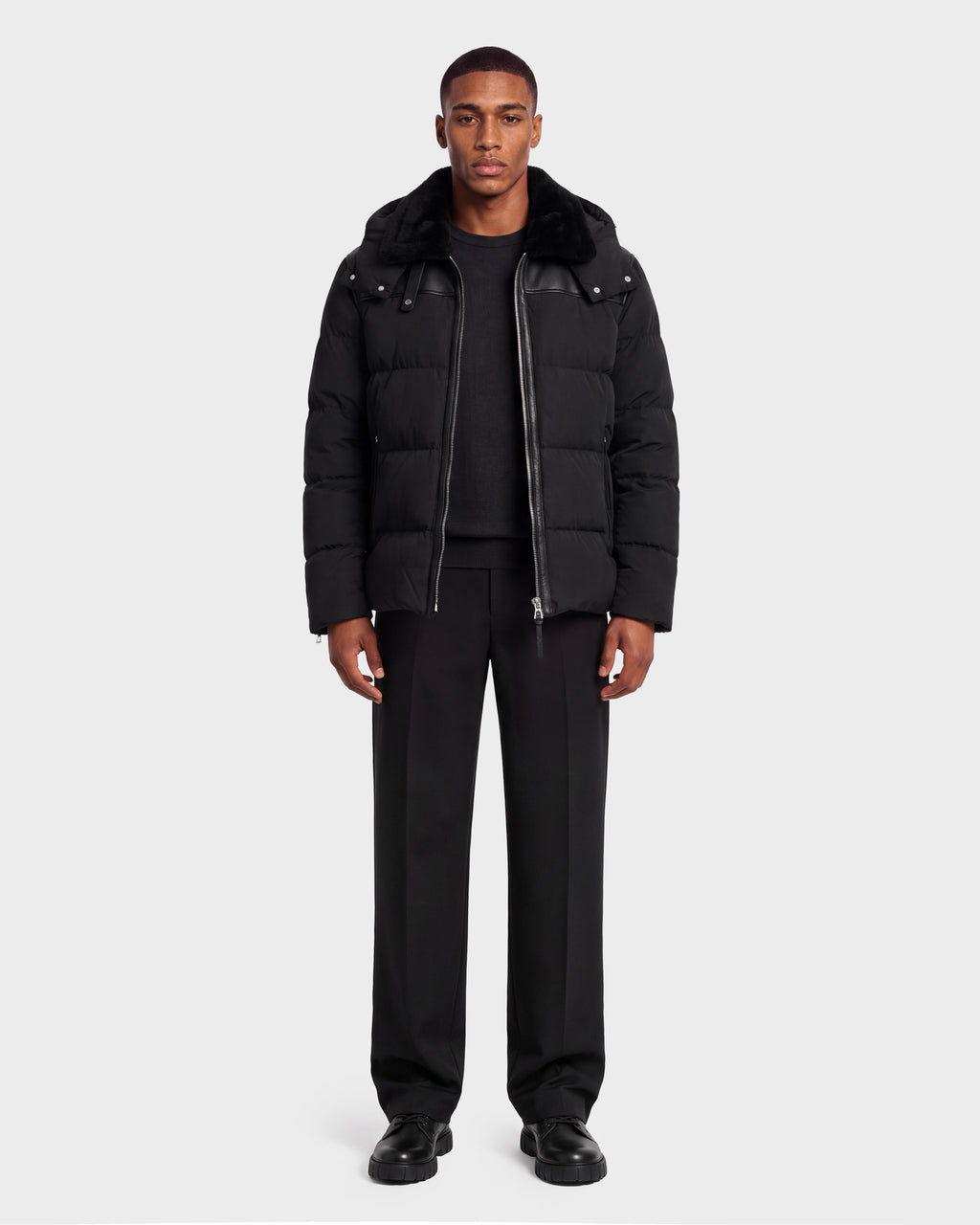 A man wearing a black Rudsak SIMON PX - 6126503 Black quilted puffer jacket with a fur-trimmed hood over a black sweater and trousers.