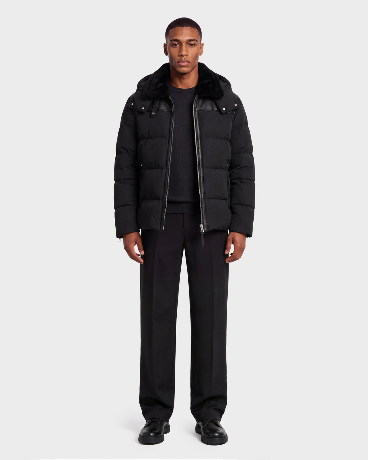 A man wearing a black Rudsak SIMON PX - 6126503 Black quilted puffer jacket with a fur-trimmed hood over a black sweater and trousers.