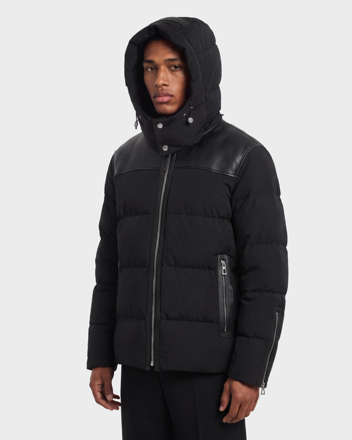 A person wears a black Rudsak SIMON PX - 6126503 Black quilted puffer jacket featuring a hood, leather-look shoulder panels, and zippered pockets.