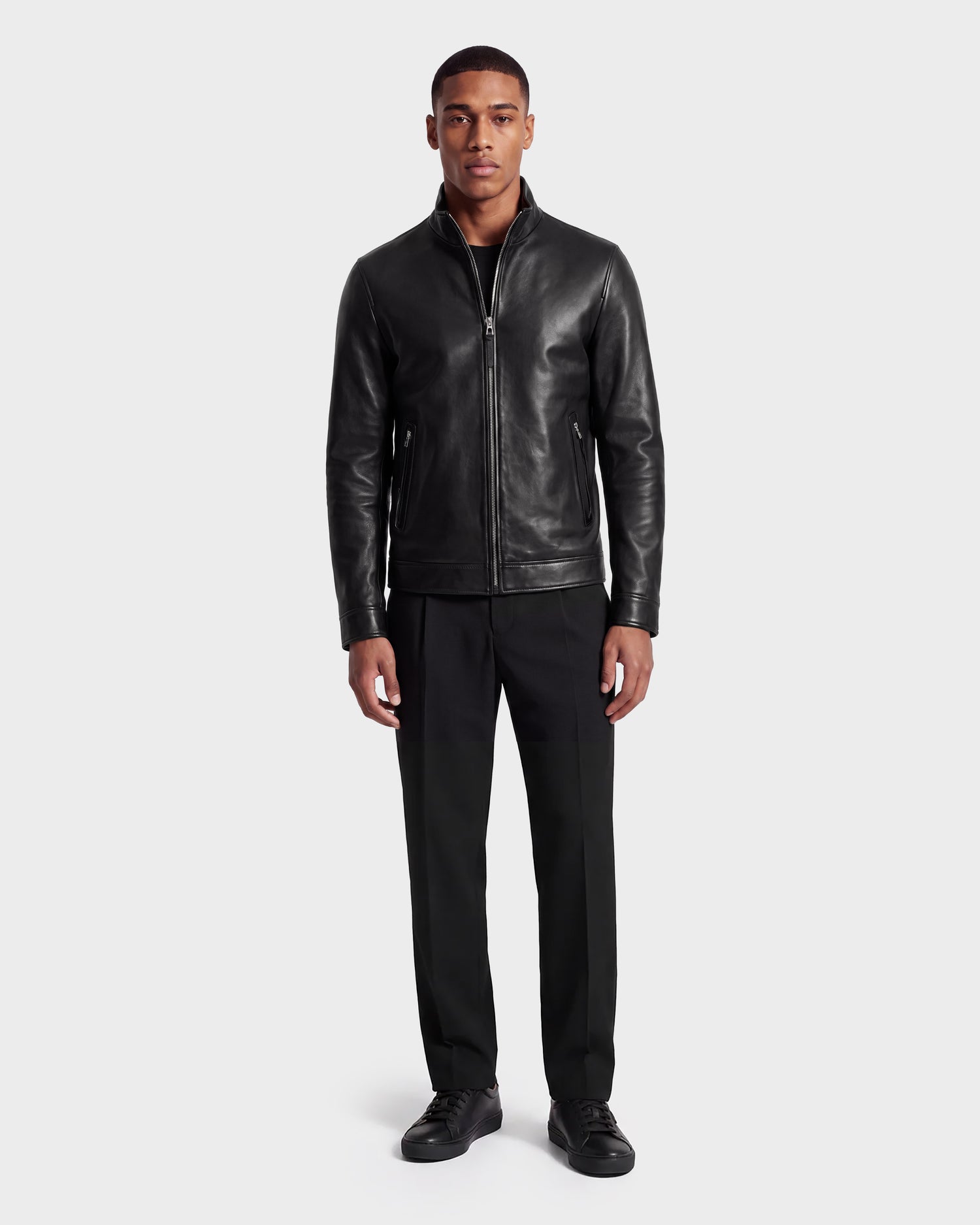 A man stands facing forward, wearing the STEVEN - 6124541 Black leather jacket by Rudsak, paired with a black shirt, black pants, and black shoes, against a plain light background.