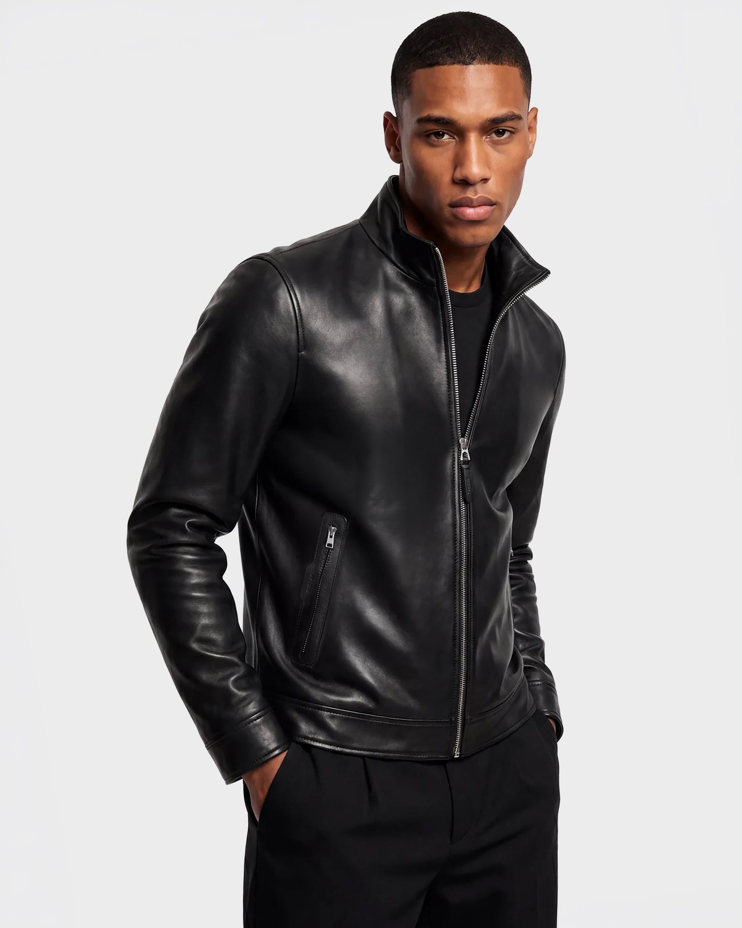 A man wearing the Rudsak STEVEN - 6124541 Black leather jacket and black pants stands with his hands in his pockets against a plain light background.