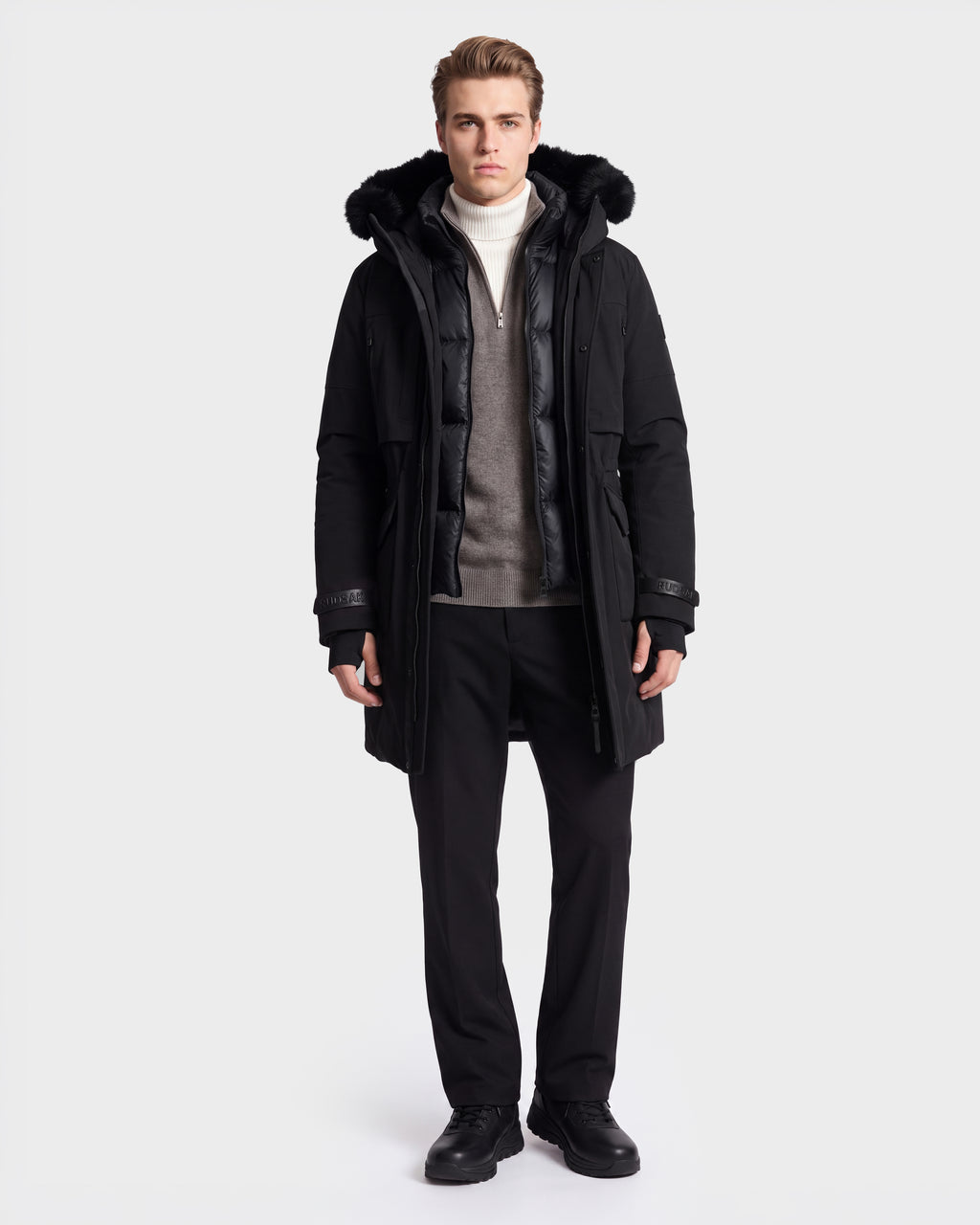 A person stands facing forward against a plain white background, wearing the Rudsak TROY - 6126504 Black coat with a fur-lined hood over a gray jacket, white turtleneck, black pants, and black shoes.
