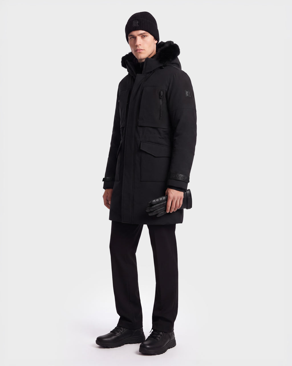 A man stands wearing the Rudsak TROY - 6126504 Black winter parka, paired with black pants, boots, a knit hat, and holding black gloves. The background is plain white.
