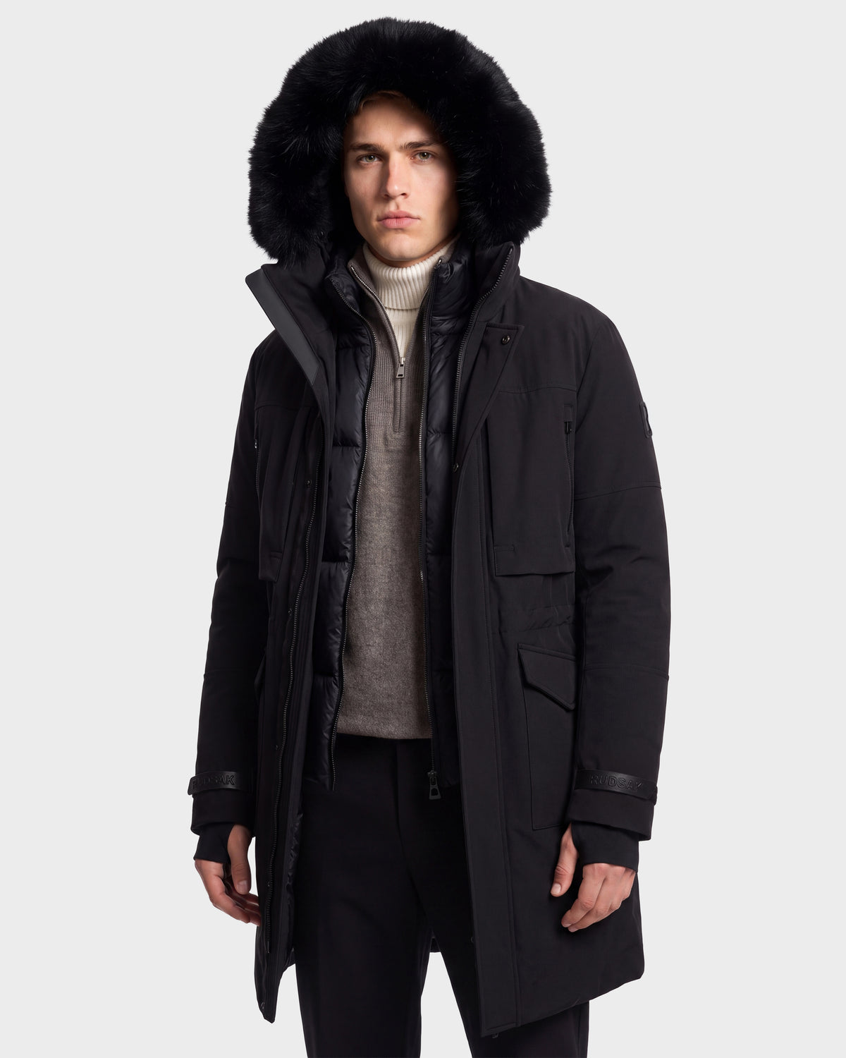 A person wearing the Rudsak TROY - 6126504 Black winter parka, featuring a large, furry hood and an eco-conscious recycled nylon shell, is layered over a light turtleneck sweater and grey pullover, standing against a plain light background.