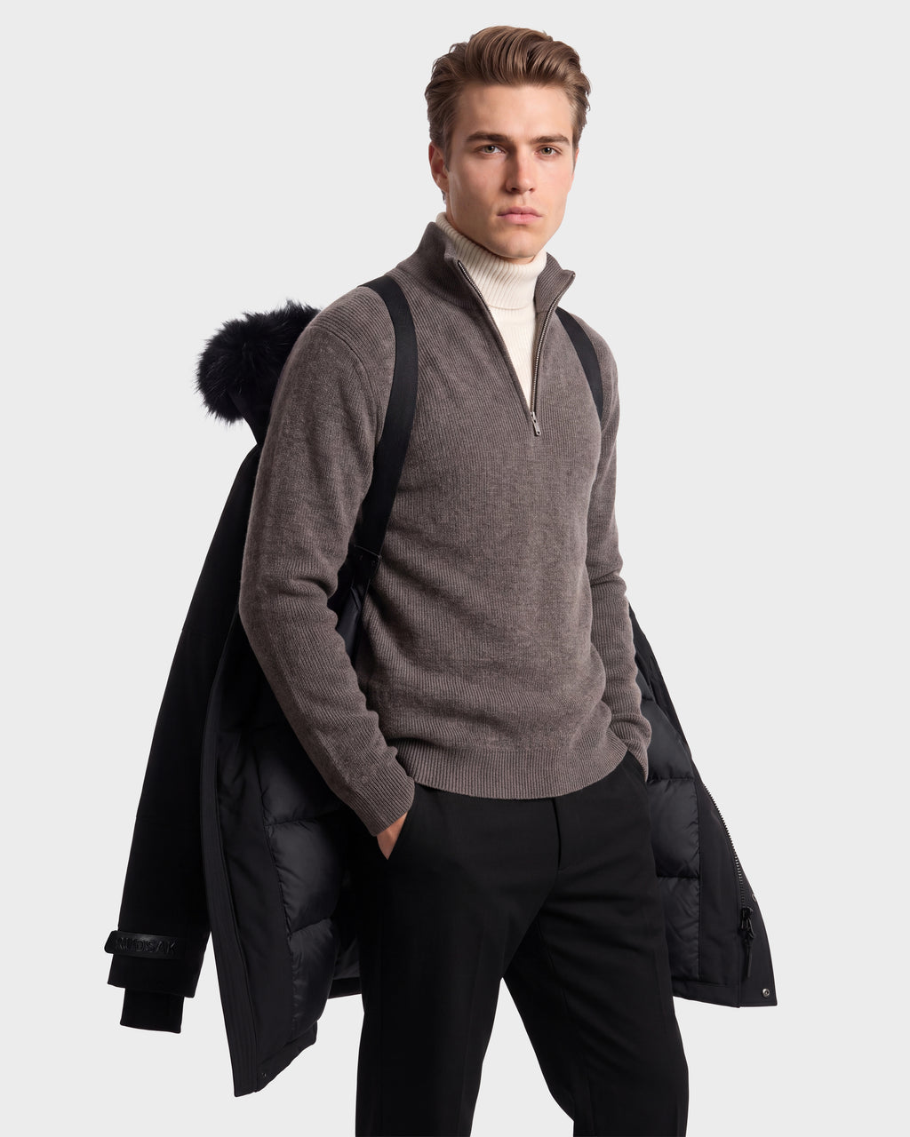 A young man stands confidently against a plain background, a Rudsak TROY - 6126504 Black coat draped over his shoulders atop a taupe quarter-zip sweater and white turtleneck, with a backpack at his side.