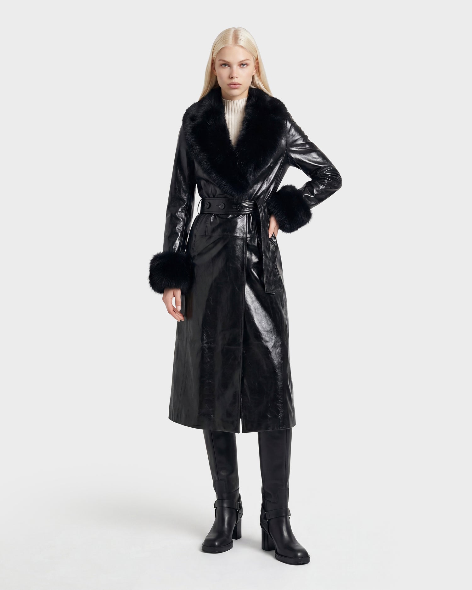 A woman stands against a plain white background, wearing the Rudsak VIVIANNE - 8126933 Black: a long black faux leather coat featuring a fur collar and cuffs, paired with a white turtleneck sweater, black knee-high boots, and a belt. This stylish look is available for pre-order with expected delivery in August 2026.