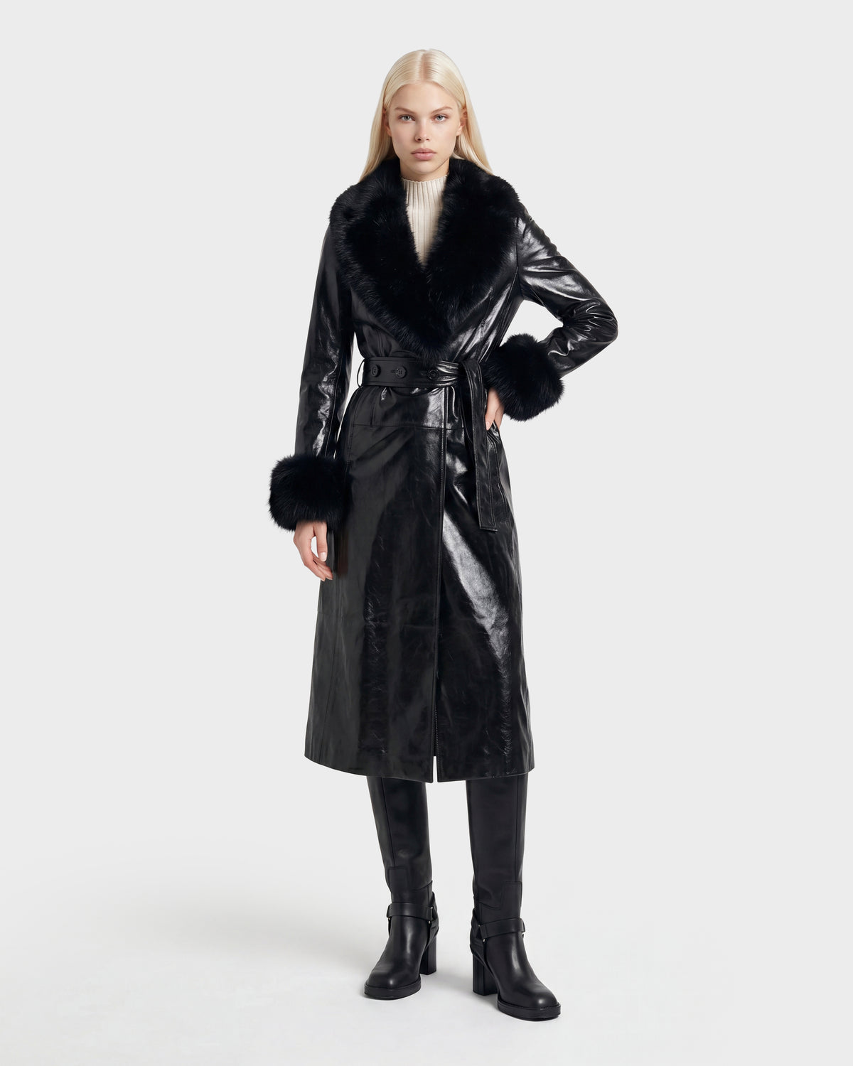 A woman with long blonde hair wears the VIVIANNE - 8126933 Black belted coat by Rudsak, featuring a shiny finish, faux fur collar and cuffs, paired with black knee-high boots and a white turtleneck. She stands against a plain light background. This stylish look is now available for pre-order, with expected delivery beginning August 17, 2026.