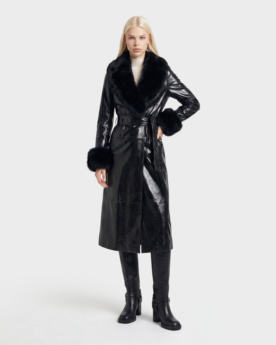 A woman stands against a plain white background, wearing the Rudsak VIVIANNE - 8126933 Black: a long black faux leather coat featuring a fur collar and cuffs, paired with a white turtleneck sweater, black knee-high boots, and a belt. This stylish look is available for pre-order with expected delivery in August 2026.