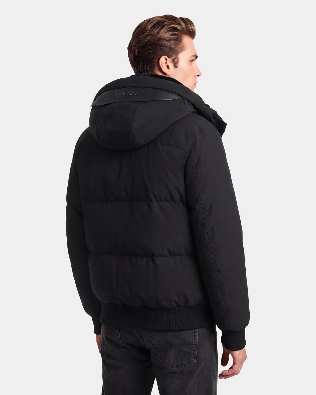 A man with short brown hair is wearing the Rudsak WILL - 6126561 Black puffer jacket with a hood and black jeans, standing and facing away from the camera against a plain white background.