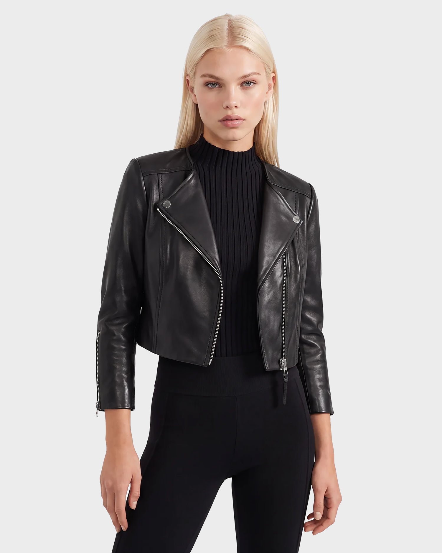 A woman with straight blonde hair models the Rudsak WINNI - 8124053 Black, a women's cropped lamb leather biker jacket, layered over a ribbed black top and black pants as she stands against a plain light gray background.