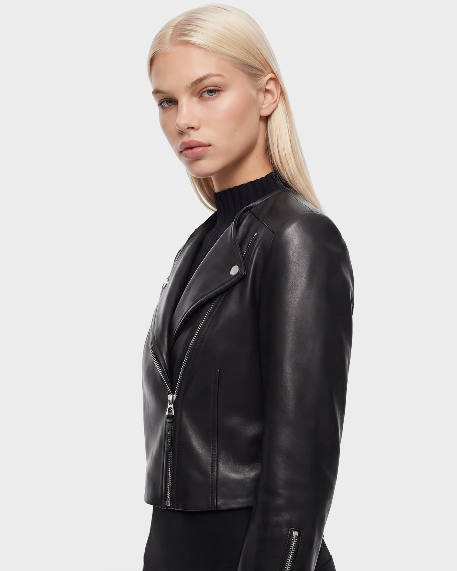 A person with straight blonde hair wears the Rudsak WINNI - 8124053 Black cropped biker jacket in black lamb leather over a black top, standing against a plain light background and looking toward the camera with a neutral expression—a timeless look in women's fashion.