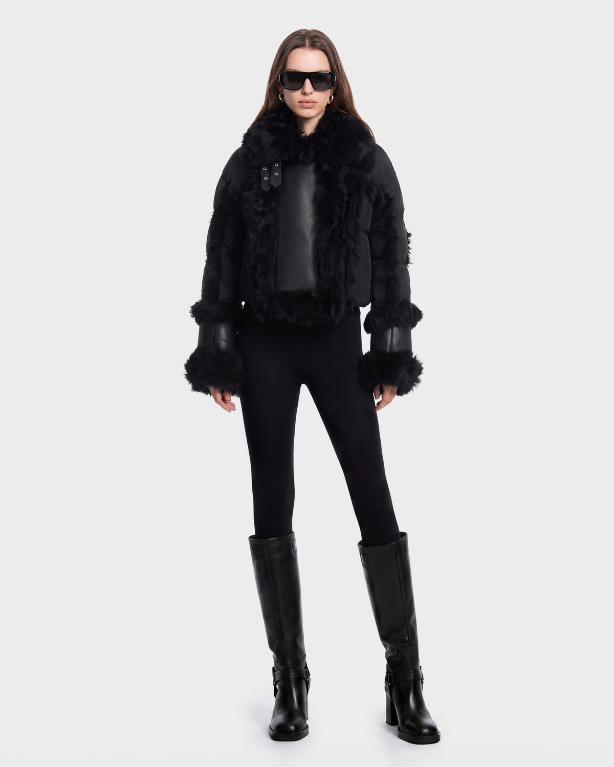 A person stands facing forward against a plain white background wearing the ZELAIA - 8125577 Black cropped puffer with faux-fur detailing by Rudsak, paired with black sunglasses, black leggings, and knee-high black boots.