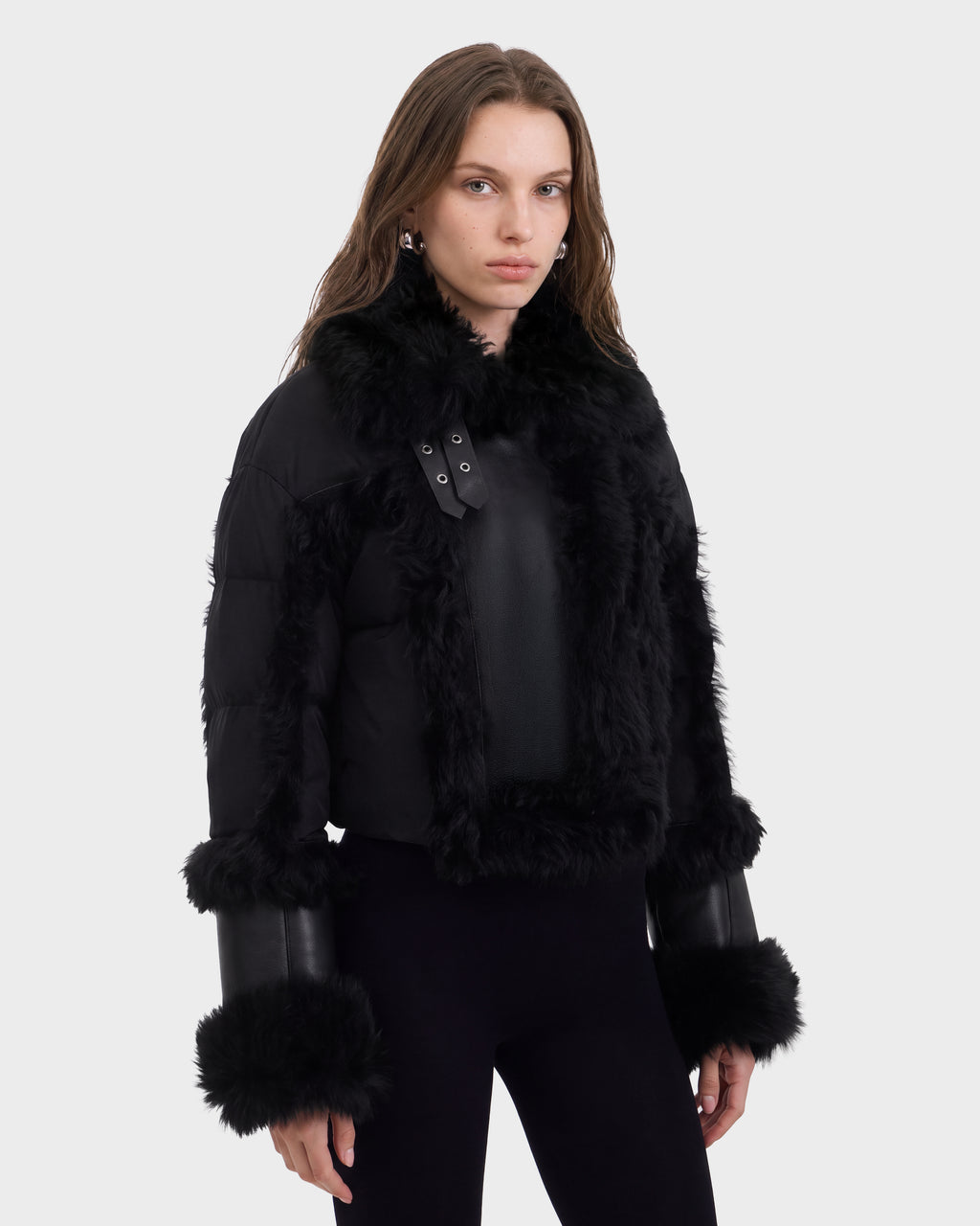 A woman with long brown hair wears the Rudsak ZELAIA - 8125577 Black jacket featuring luxurious faux-fur detailing, paired with black pants and silver earrings, standing against a plain white background.