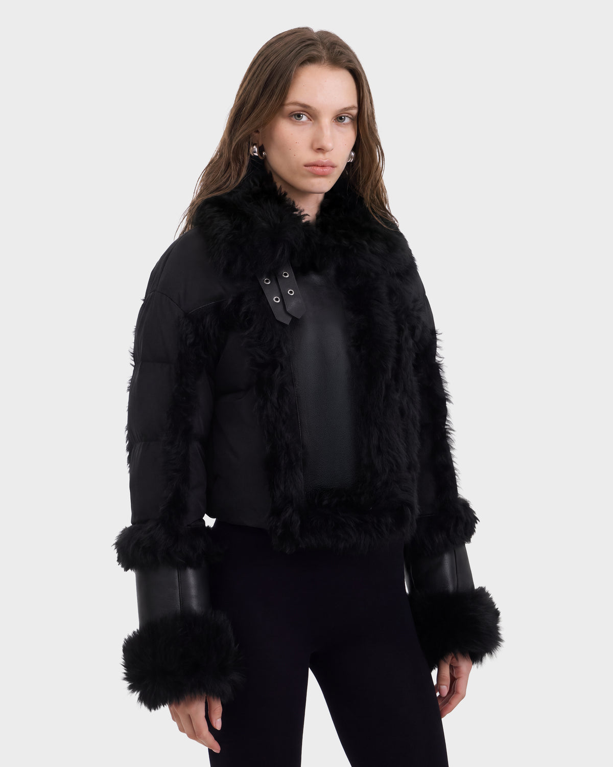 A woman with long brown hair wears the Rudsak ZELAIA - 8125577 Black jacket featuring luxurious faux-fur detailing, paired with black pants and silver earrings, standing against a plain white background.