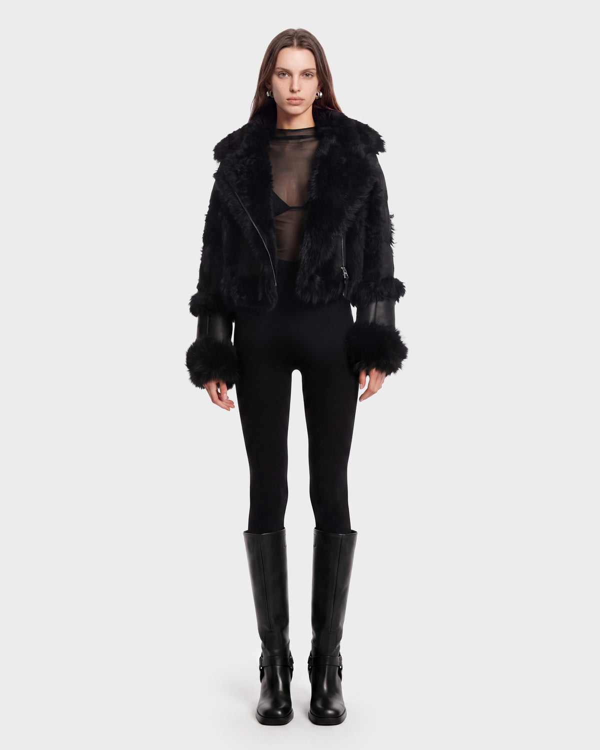 A woman stands facing forward against a plain white background, wearing the Rudsak ZELAIA - 8125577 Black cropped puffer with faux-fur detailing, styled over a sheer black top, black pants, and tall black boots.