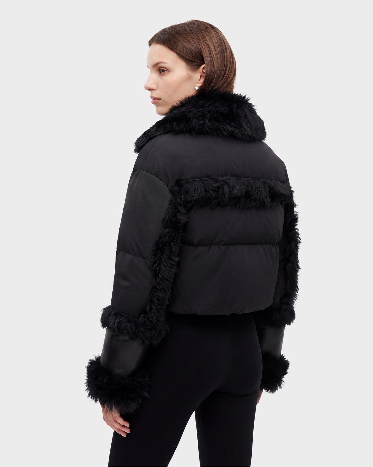 A woman stands facing away, looking over her shoulder against a plain light background, wearing the Rudsak ZELAIA - 8125577 Black cropped puffer jacket with faux-fur detailing and black pants.