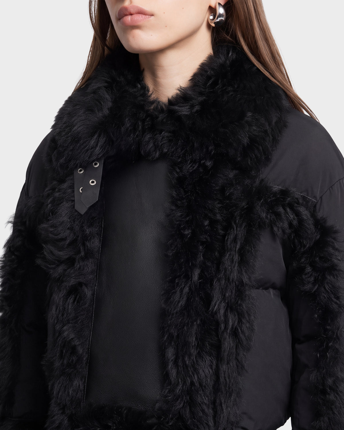 A woman wearing the Rudsak ZELAIA - 8125577 Black coat, featuring large, fluffy faux-fur detailing on the collar and trim, along with silver earrings and straight brown hair, shown from the shoulders up against a plain background.