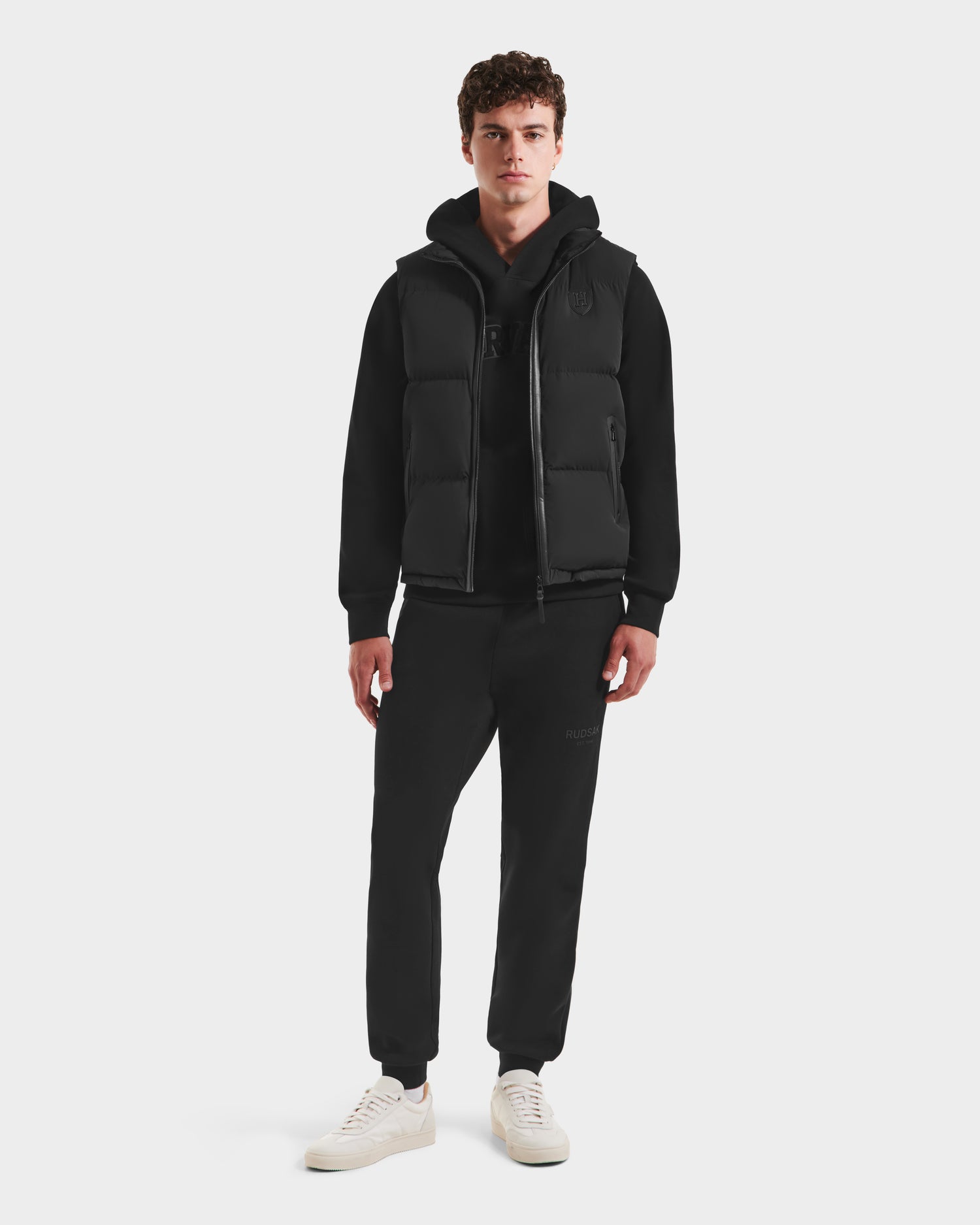 A young man stands facing forward in Rudsak’s FREDDY HV - H125510 Black hooded sweatshirt, a limited-edition quilted down puffer vest, black jogger pants, and white sneakers against a plain light background.