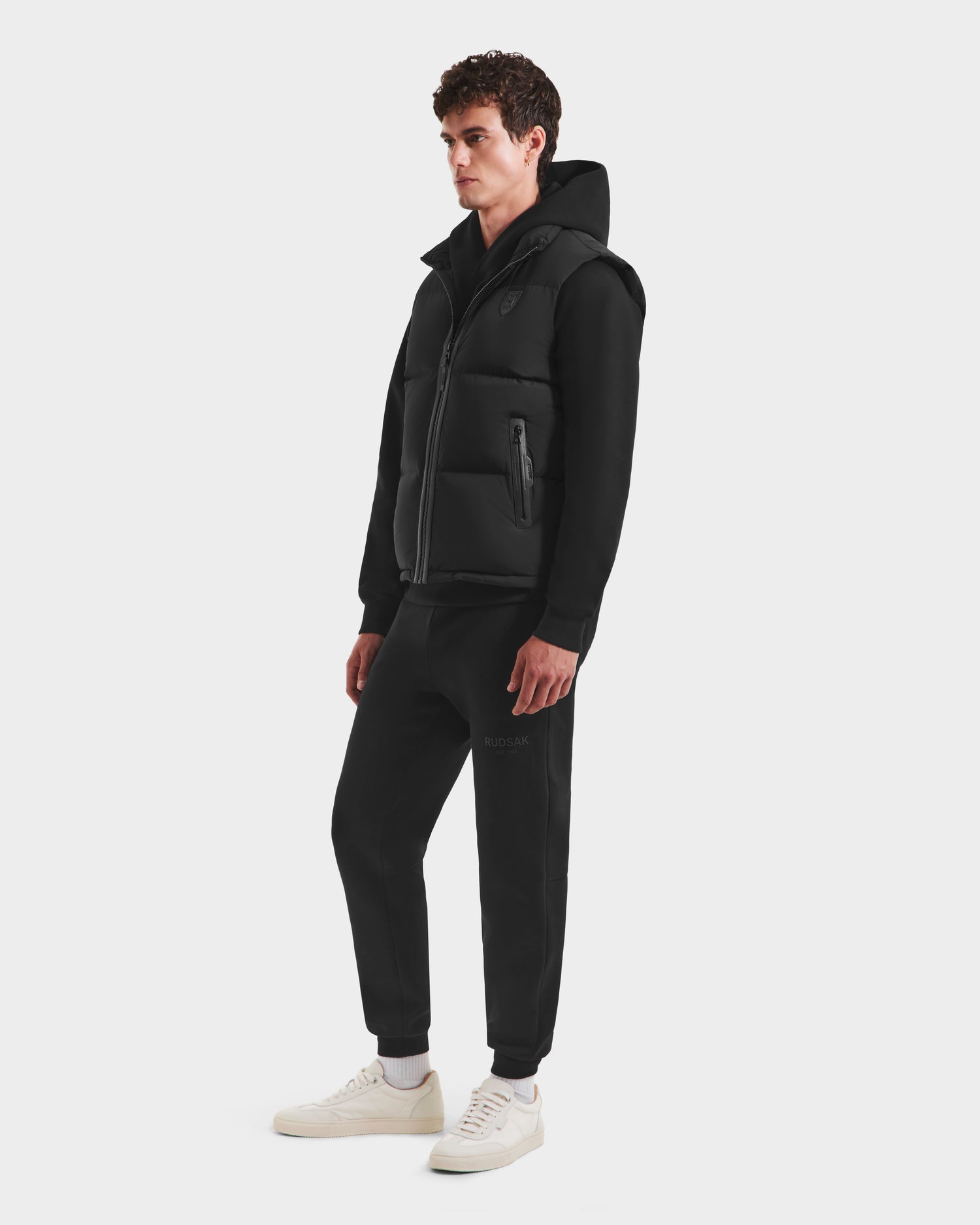 A young man wears the FREDDY HV - H125510 Black quilted down puffer vest by Rudsak over a black sweatshirt and joggers with white sneakers, standing against a plain white background.
