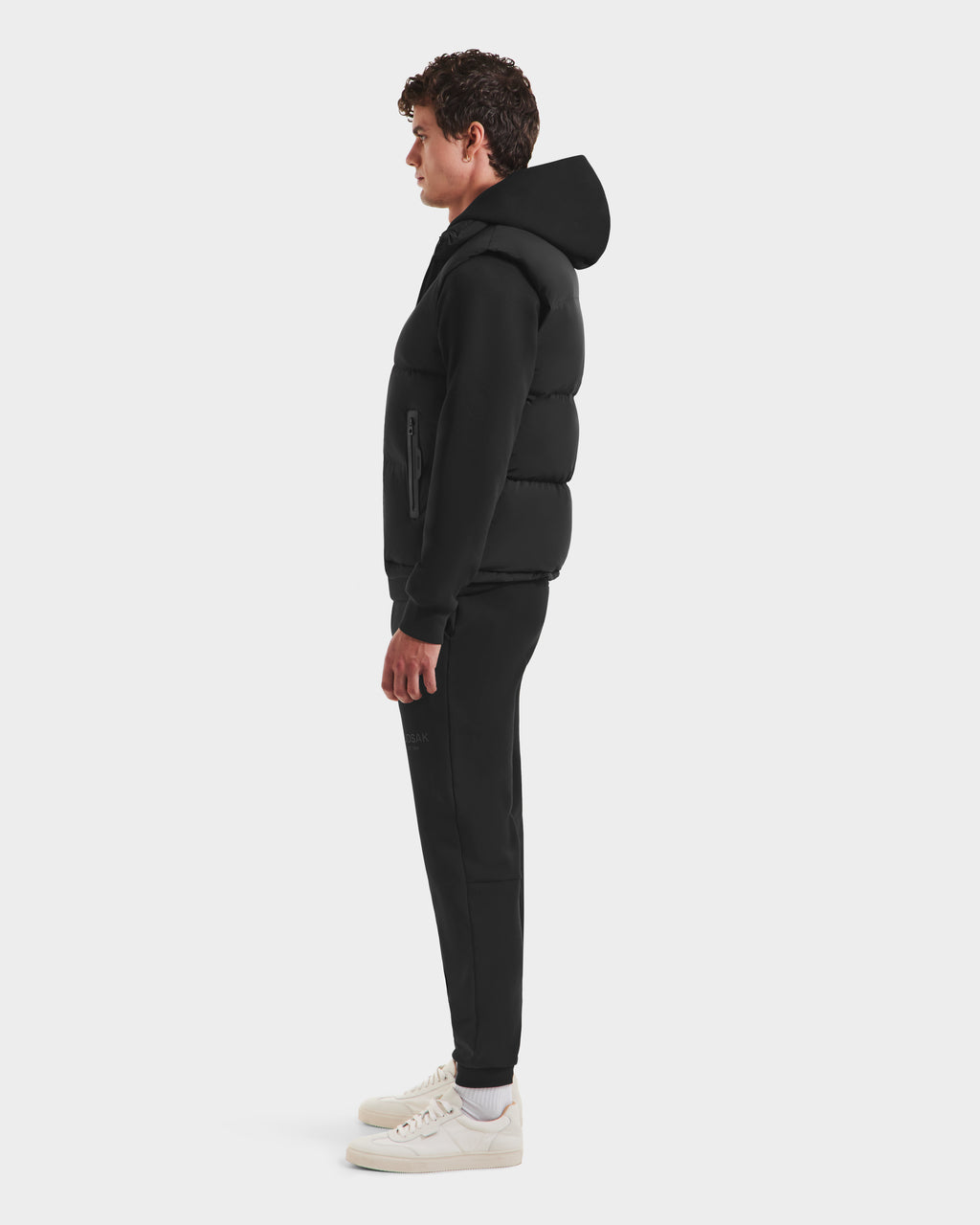 A man in profile faces right, wearing the Rudsak FREDDY HV - H125510 Black quilted down puffer jacket with a hood, black jogger pants, and white sneakers, standing against a plain white background.