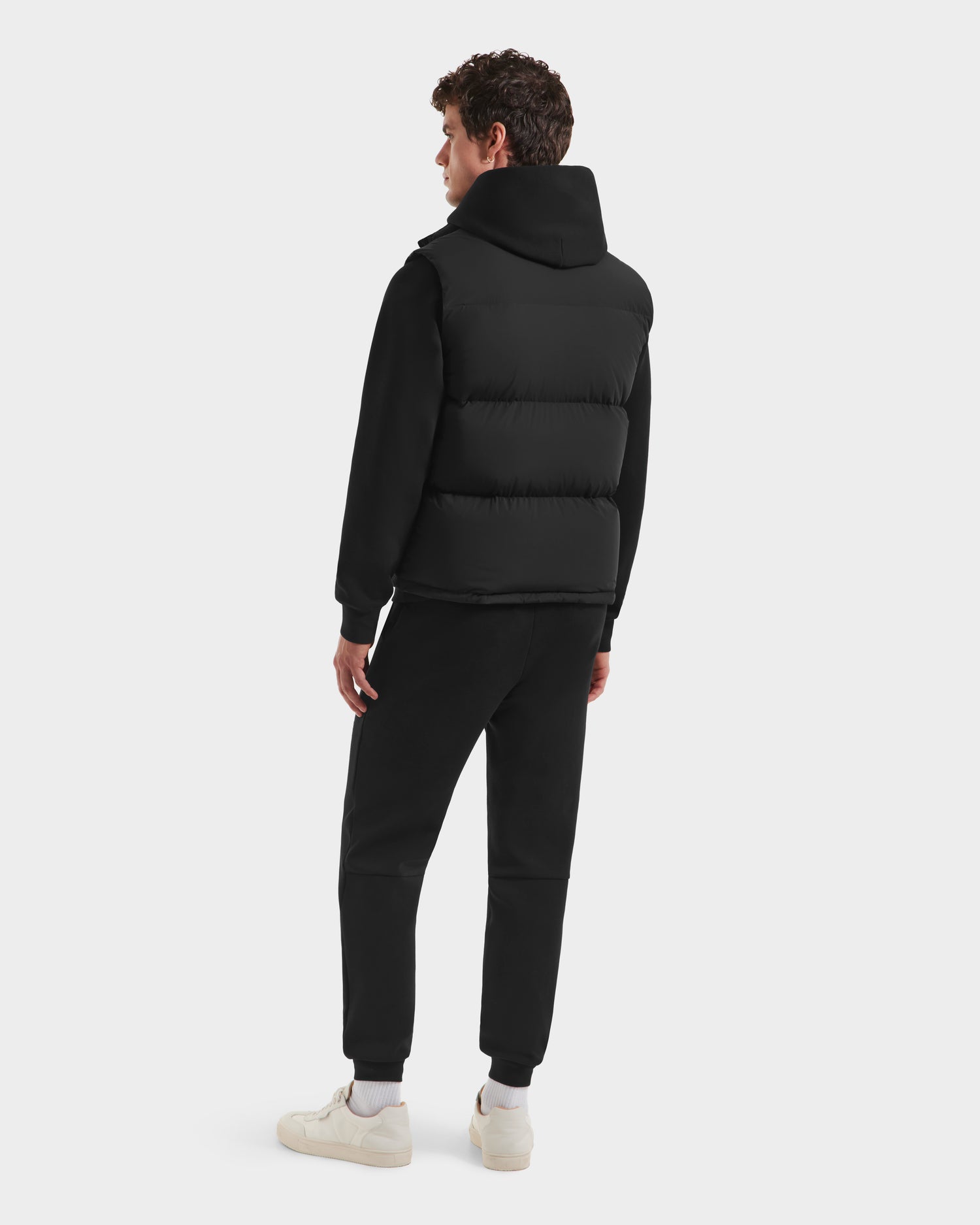 A person faces away, wearing the limited-edition Rudsak FREDDY HV - H125510 Black quilted down puffer vest over a black sweatshirt and pants, with white sneakers, against a plain light background.