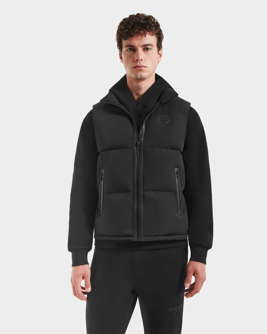 A young man with curly brown hair wears the Rudsak FREDDY HV - H125510 Black quilted down puffer vest over a black hoodie and sweatpants, standing against a plain white background.