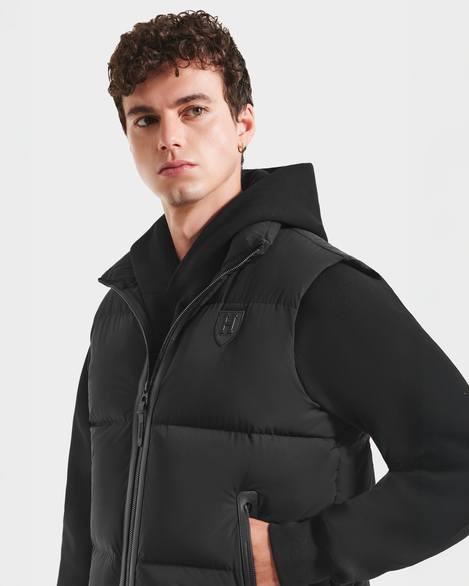 A young man with curly brown hair models the Rudsak FREDDY HV - H125510 Black hooded sweatshirt and limited-edition Harvard x RUDSAK sleeveless quilted down puffer vest, standing with one hand in his pocket against a plain background.
