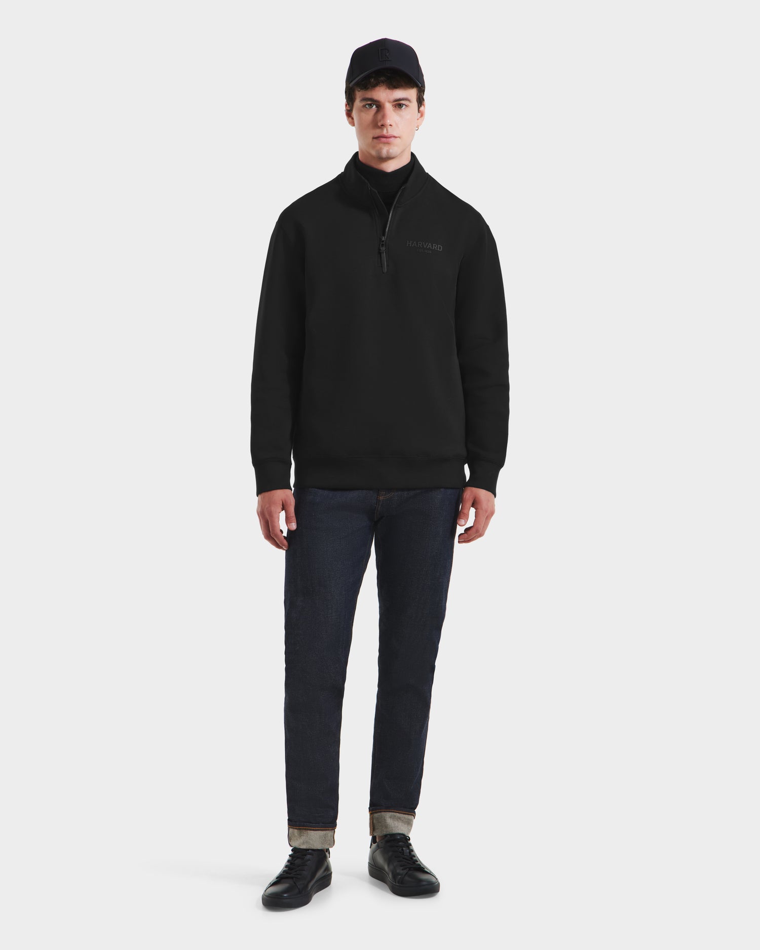A man wearing the Rudsak FREMONT HV - H525501 Black limited-edition pullover, made from a premium cotton blend, paired with dark rolled-cuff jeans and black sneakers stands facing forward against a plain white background.
