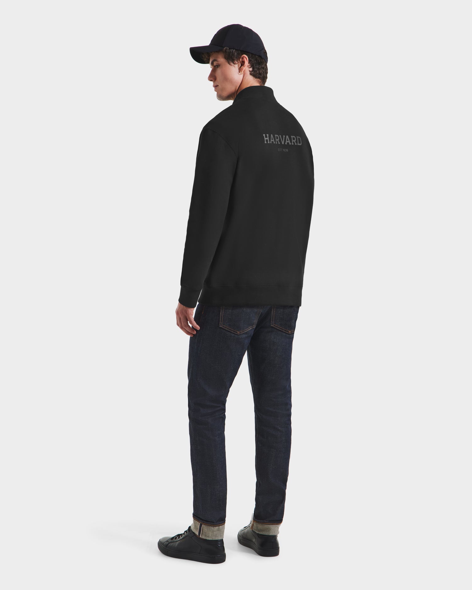A man wearing the Rudsak FREMONT HV - H525501 Black pullover, a black cap, dark jeans, and black shoes stands facing away and looks over his shoulder against a plain white background.