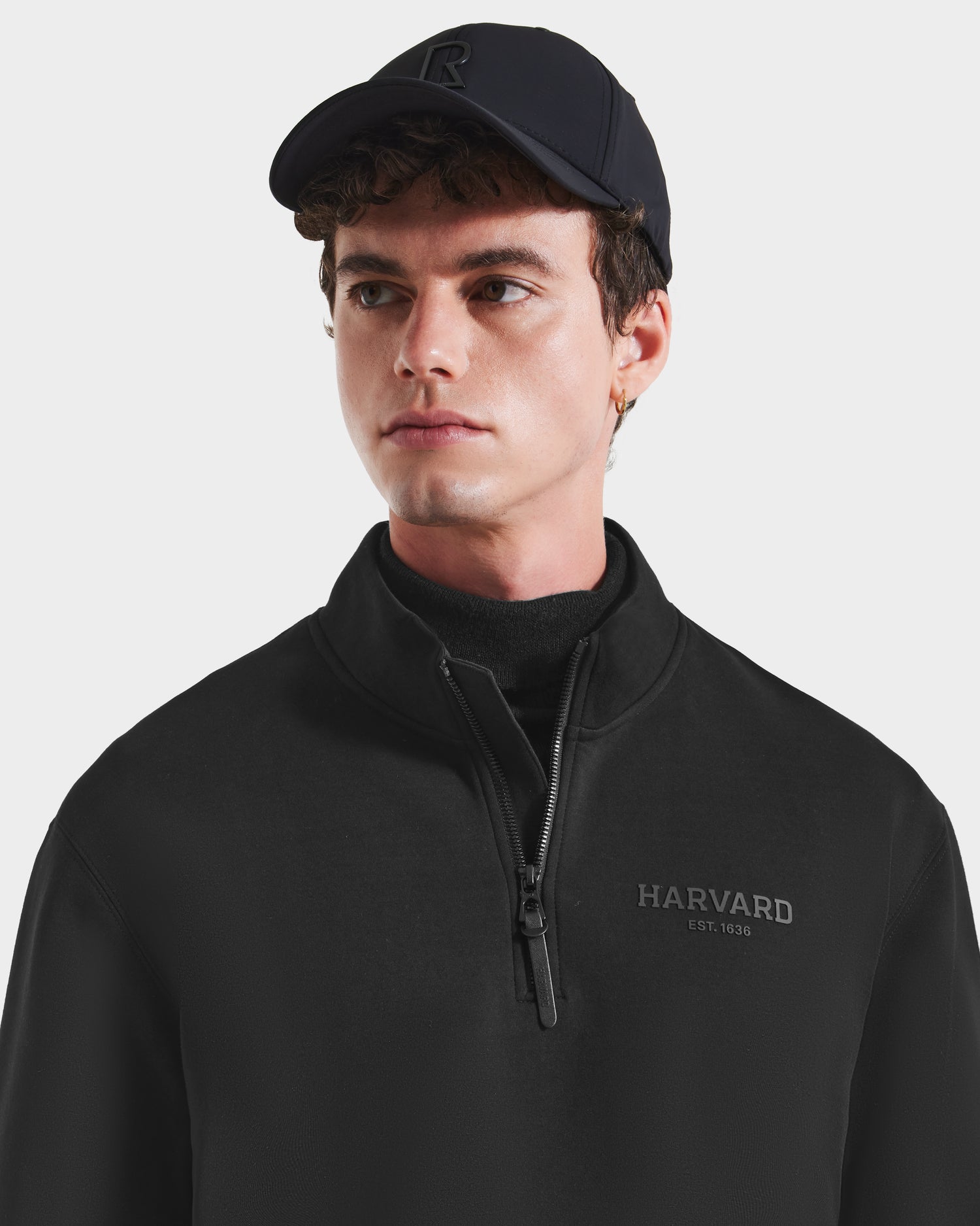 A young man wears the Rudsak FREMONT HV - H525501 Black pullover, a limited-edition cotton blend with HARVARD EST. 1636 on the front, and looks slightly to his left against a plain background.