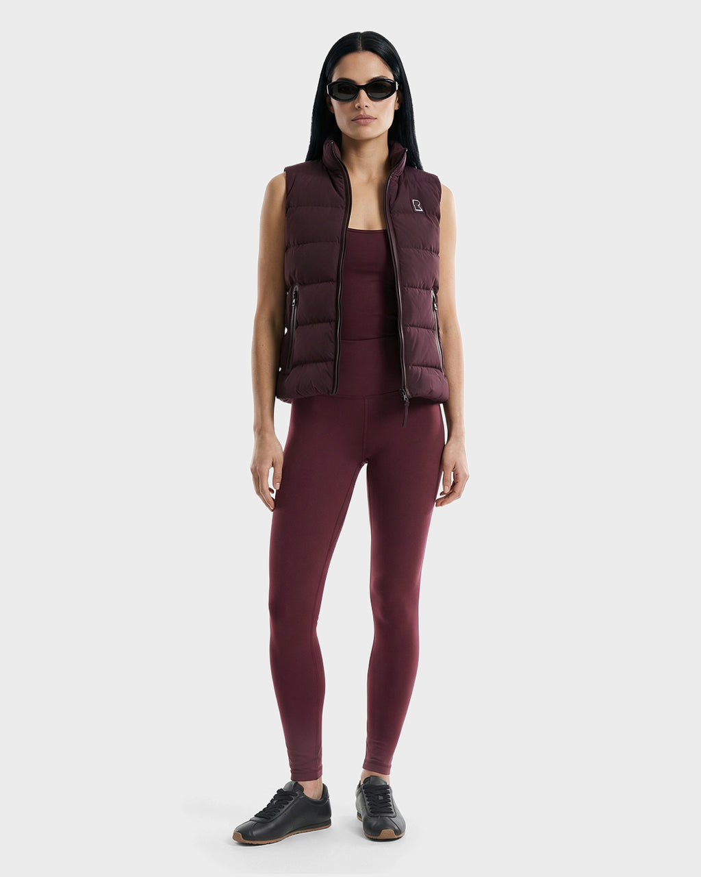 A woman with long dark hair wears black sunglasses, black sneakers, and the Rudsak GABRIELA - 8125014 Bordeaux women's down puffer vest over a matching sleeveless top and leggings, standing against a plain light gray background.
