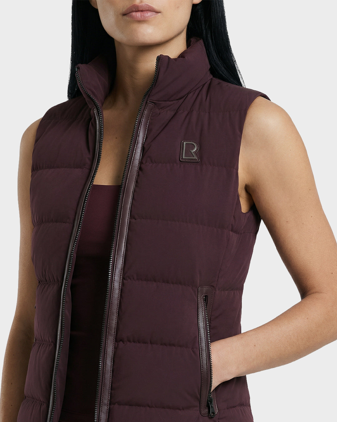 A woman with long dark hair wears the Rudsak GABRIELA - 8125014 Bordeaux women's down puffer vest with a zip front and logo patch over a matching top. Her hand rests in the pocket. The background is plain light gray.