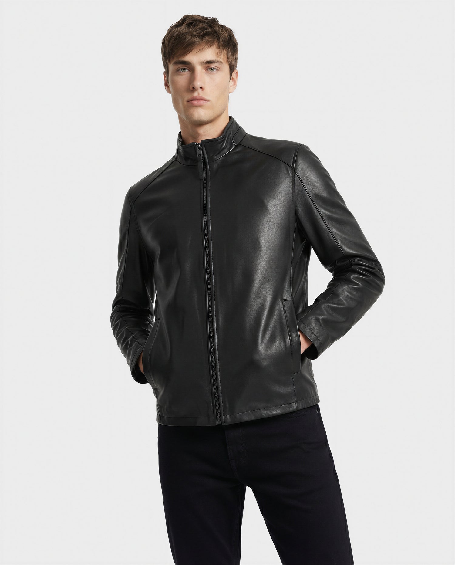 A young man with hands in pockets wears the Rudsak GARY - M126061 Black leather jacket and black pants, his expression neutral, standing against a plain light background.