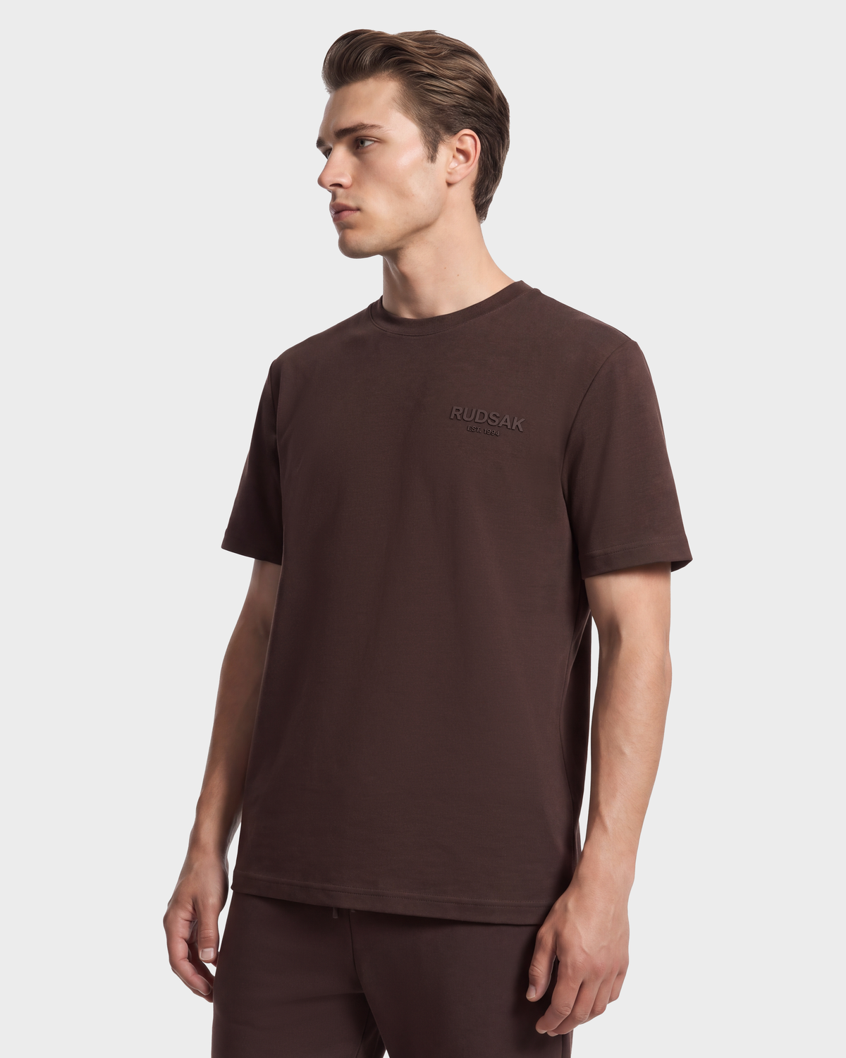 A man wearing a Rudsak GRANT - 6525036 Espresso dark brown premium cotton crewneck tee with a small logo on the chest.