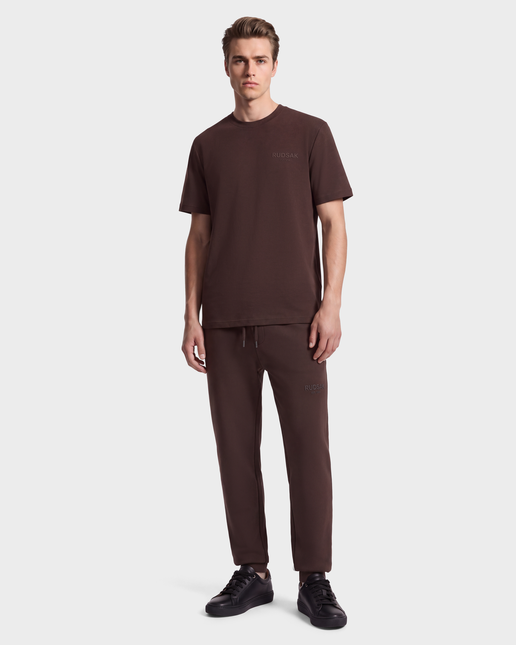 A man wearing a dark brown GRANT - 6525036 Espresso crewneck t-shirt, matching sweatpants, and black sneakers.