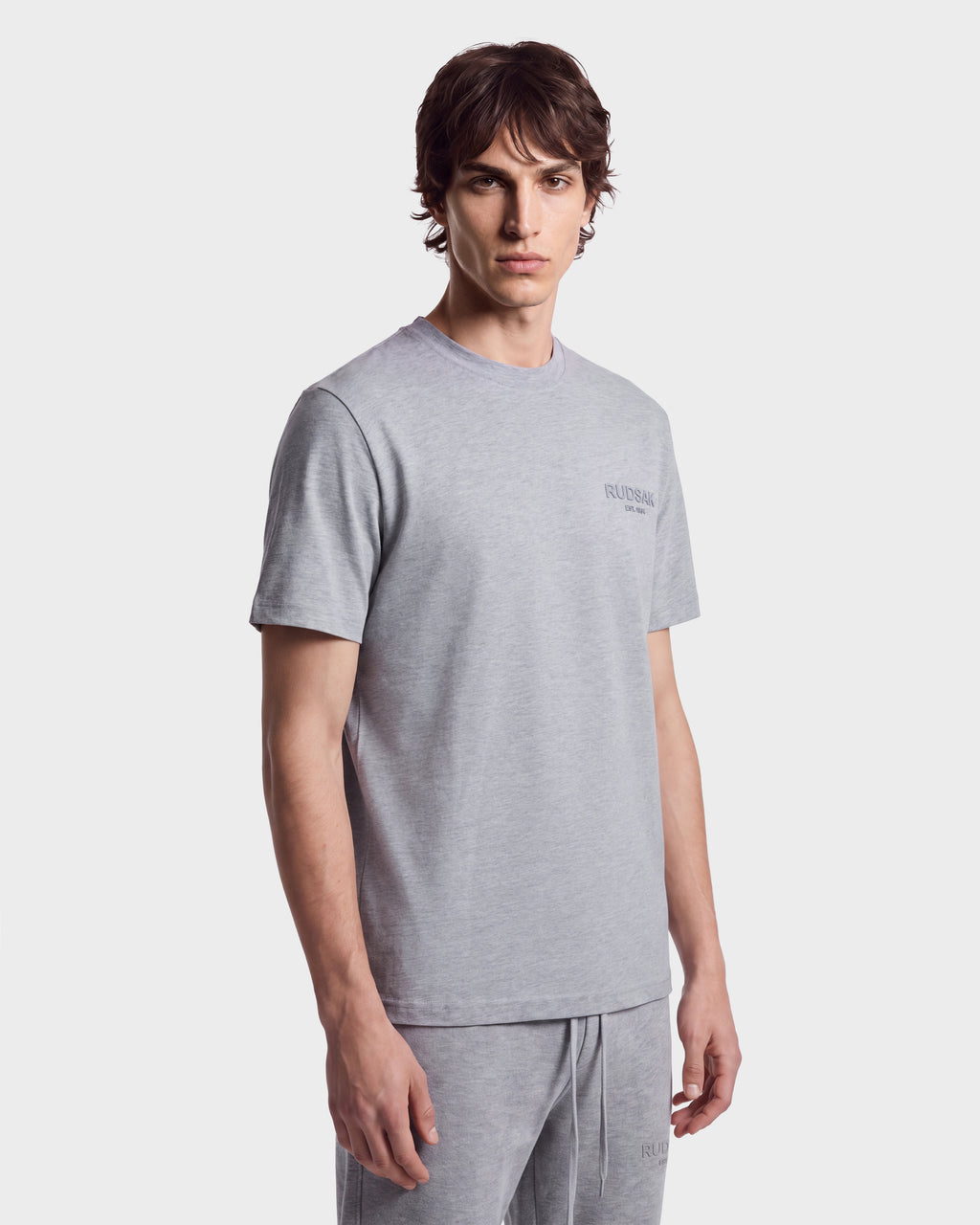 A young man with medium-length brown hair wears the Rudsak GRANT - 6525036 Heather Grey men’s tee, crafted from premium cotton with a classic crewneck design, standing against a plain white background.