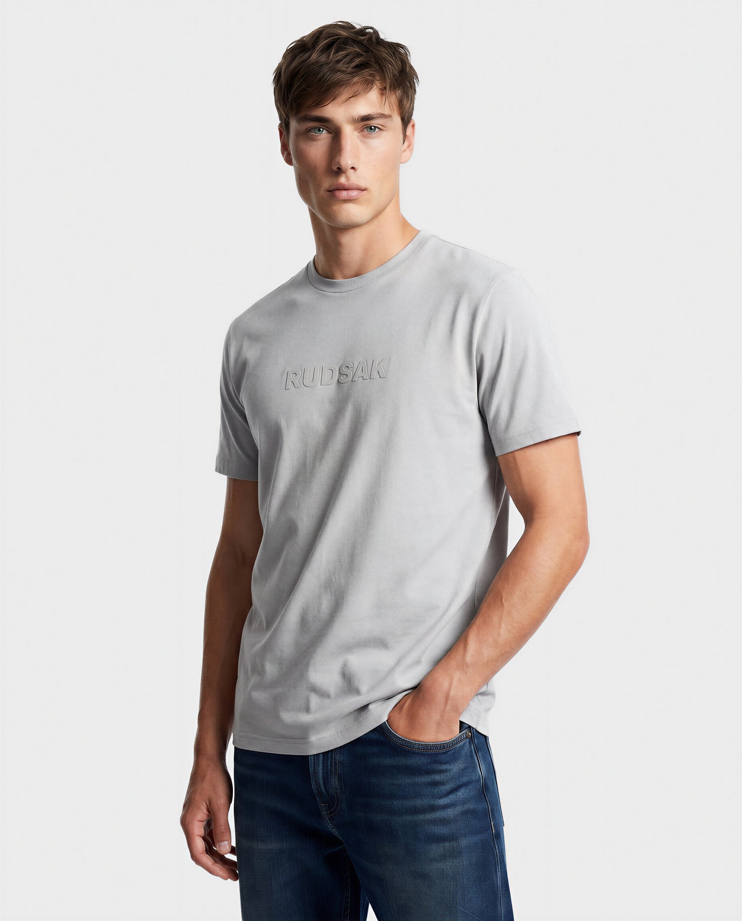 A young man stands against a plain light background wearing the Rudsak FERDI - 6522005 Mushroom t-shirt, featuring the brand’s embossed logo on the chest, with one hand in his jeans pocket.