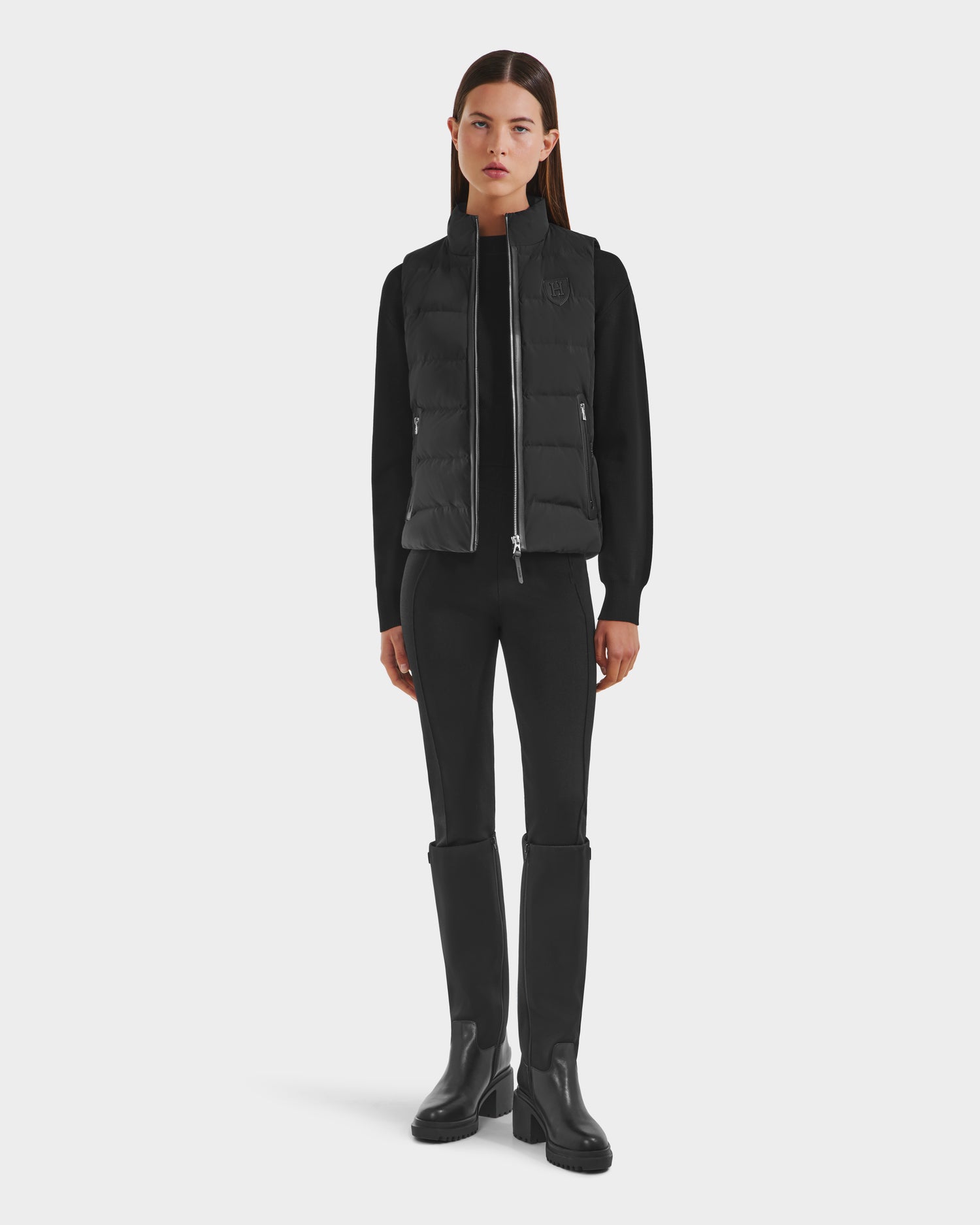 A woman faces forward against a white background, wearing the Rudsak GABRIELA HV - H125507 Black limited-edition puffer vest over a black long-sleeve top, fitted pants, and tall black boots.