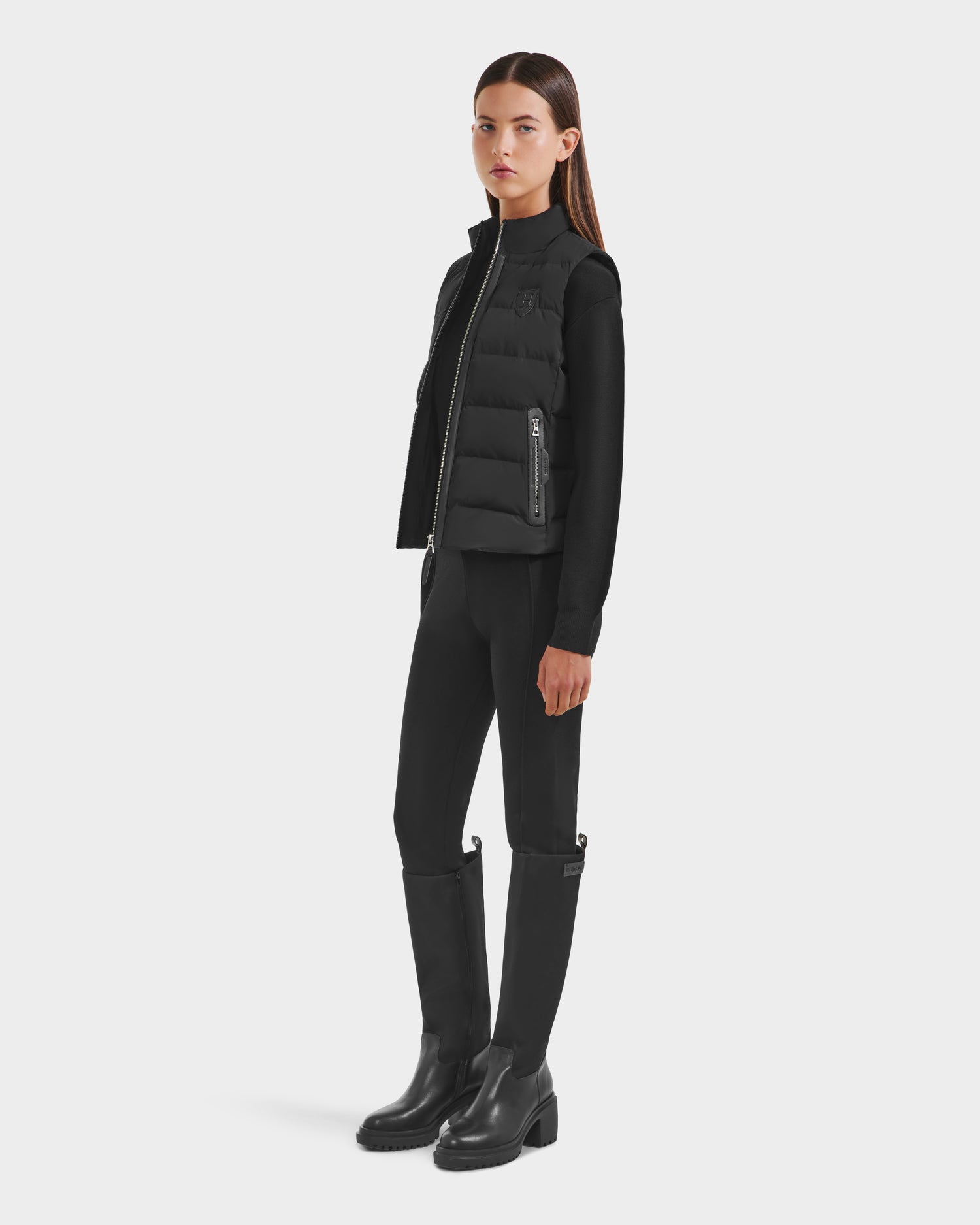A woman stands against a white background wearing the Rudsak GABRIELA HV - H125507 Black puffer vest over a black long-sleeve top, leggings, and knee-high boots. She faces slightly to the side with a neutral expression.