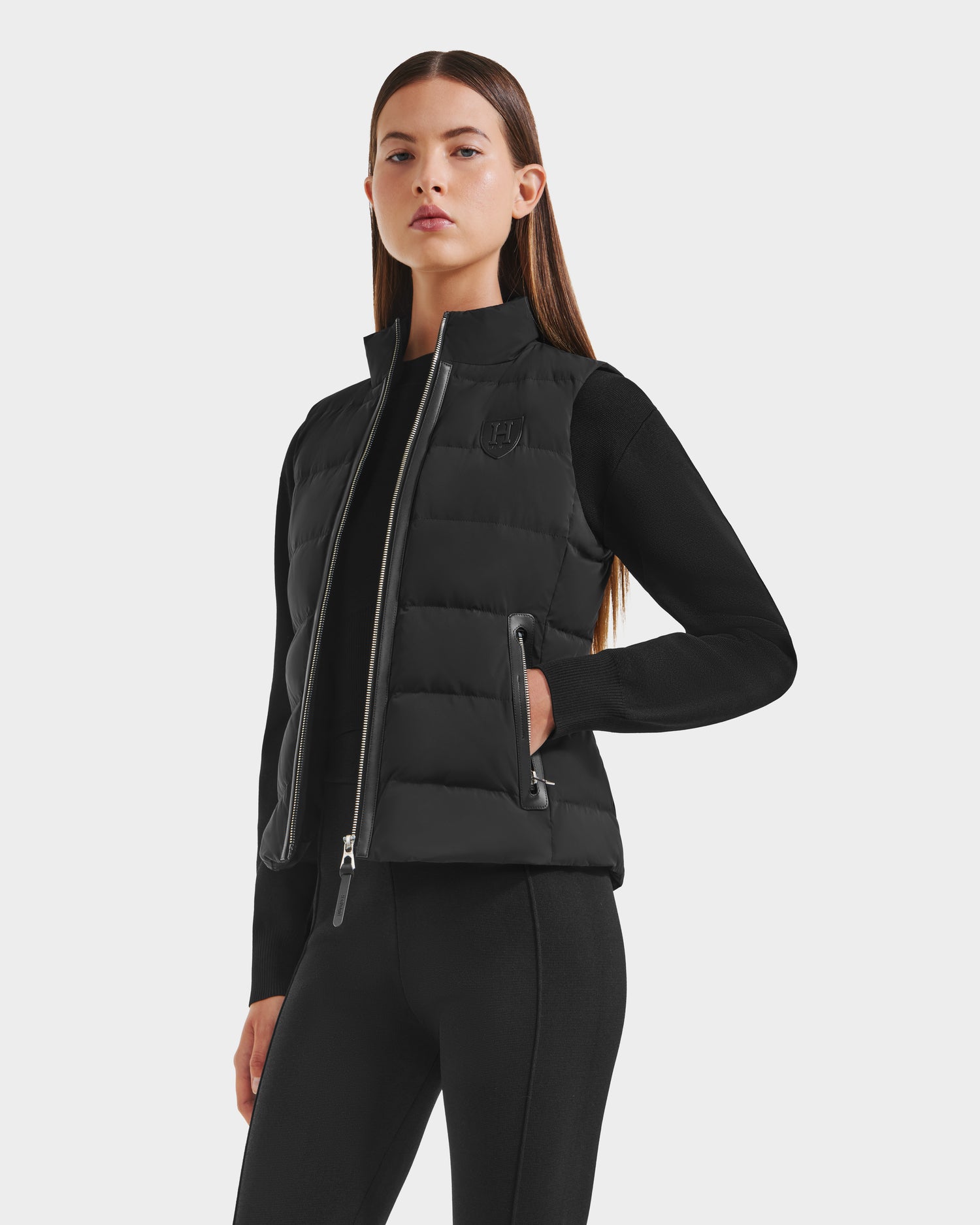 A woman with long, straight brown hair models the Rudsak GABRIELA HV - H125507 Black puffer vest over a black long-sleeve top and pants, standing with one hand in her pocket against a plain white background.